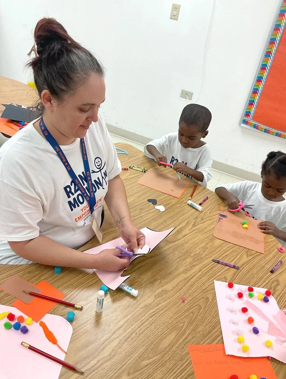 A woman and two children at a table engaged in arts and crafts activities, with colorful pom-poms, glue sticks, scissors, and paper.