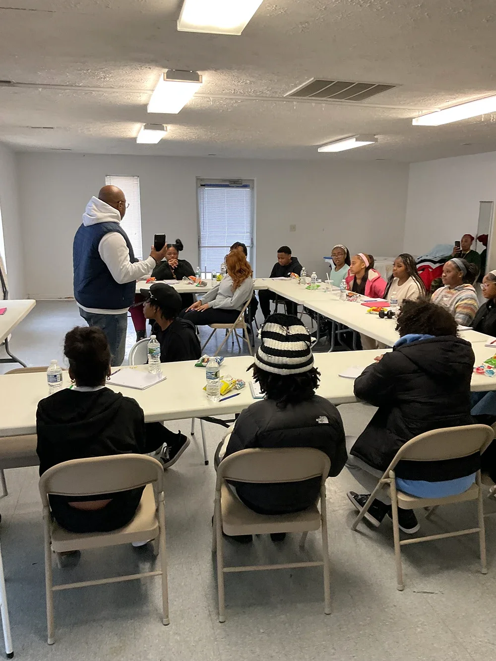 A man presenting to a diverse group of people seated at tables in a classroom or community room, with notebooks, water bottles, and snacks on the tables.