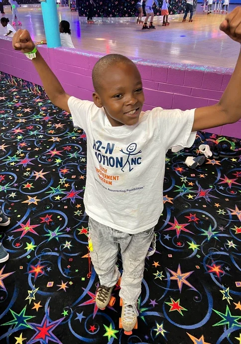 A young boy on roller skates smiling and flexing his arms in a roller skating rink with a colorful star pattern on the floor.