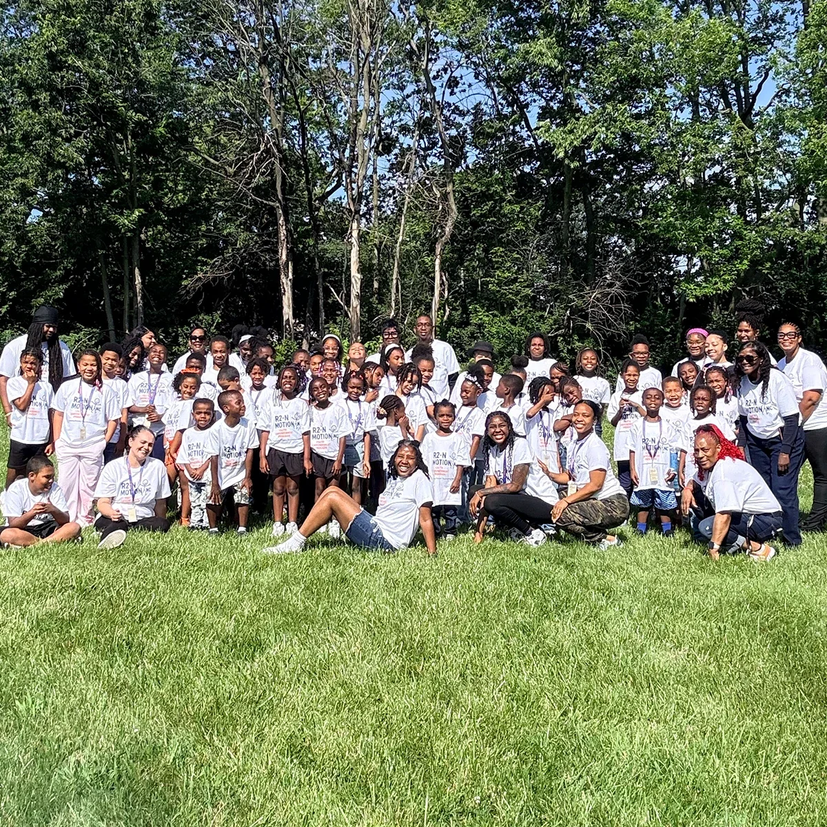 Group of children and adults outdoors on grass, posing for a photo, many wearing white t-shirts with text, surrounded by trees.