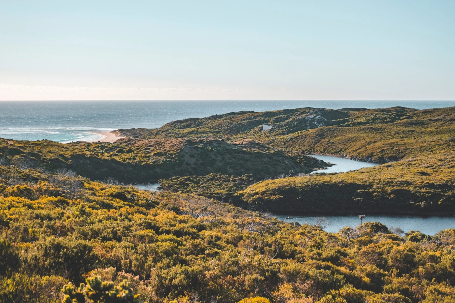 Hire Equipment Machine Western Australia South West WA - A Margaret River landscape of rolling green hills with a river flowing through them, in front of a beach with the ocean in the background under a clear sky.