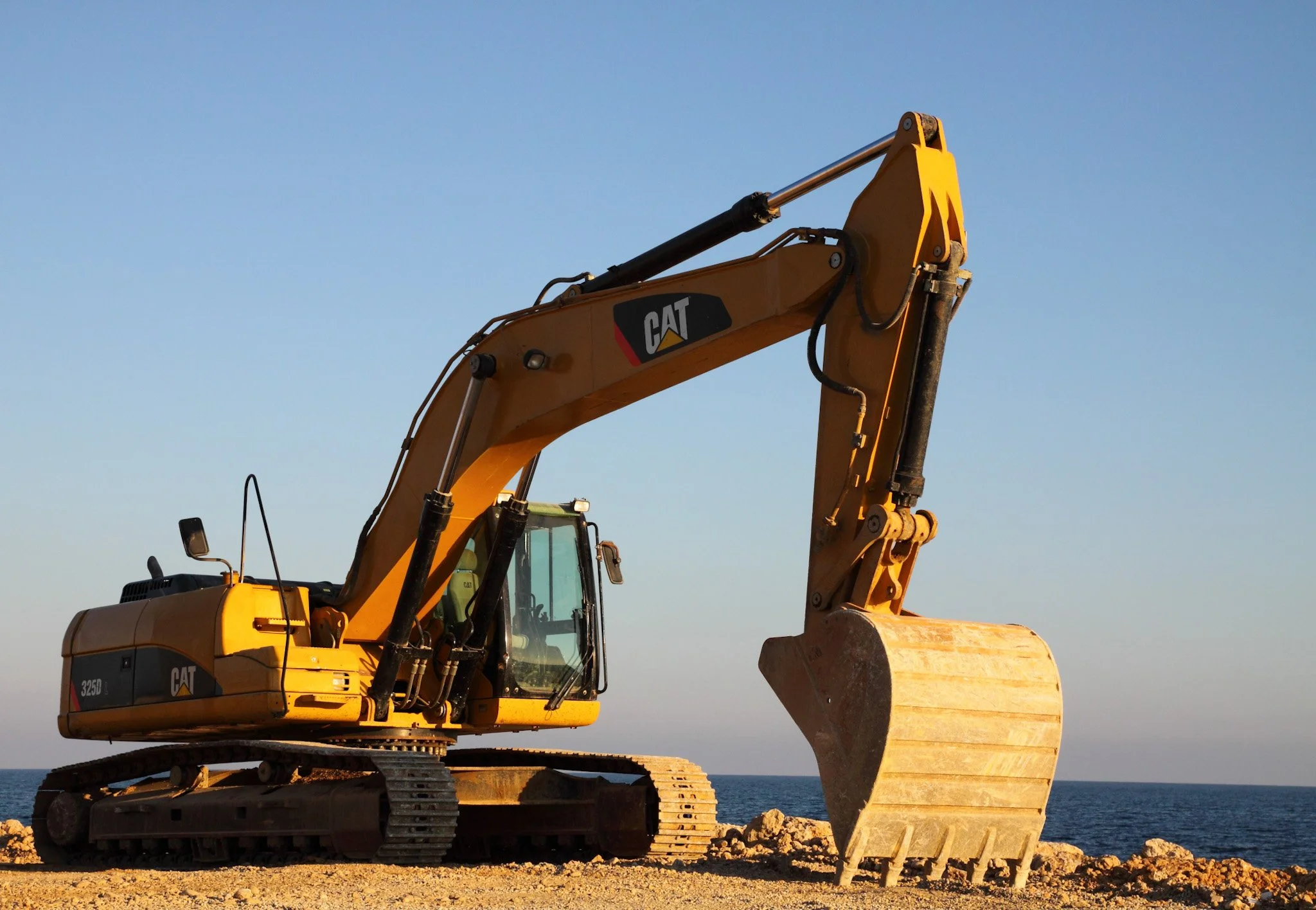 Hire Equipment Machine Western Australia South West WA - A yellow Caterpillar excavator positioned on a dirt surface near the ocean, with a clear blue sky in the background.