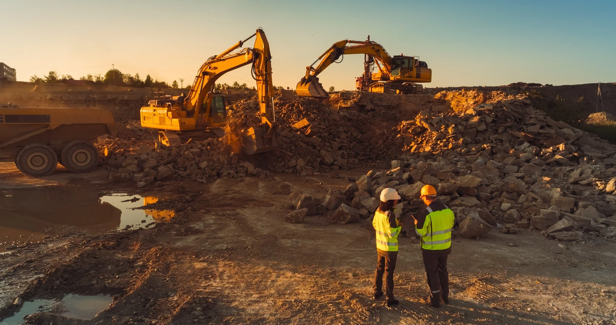 Hire Equipment Machine Western Australia South West WA - Two construction workers in safety vests and helmets reviewing plans while two excavators work on a rocky site during sunset.