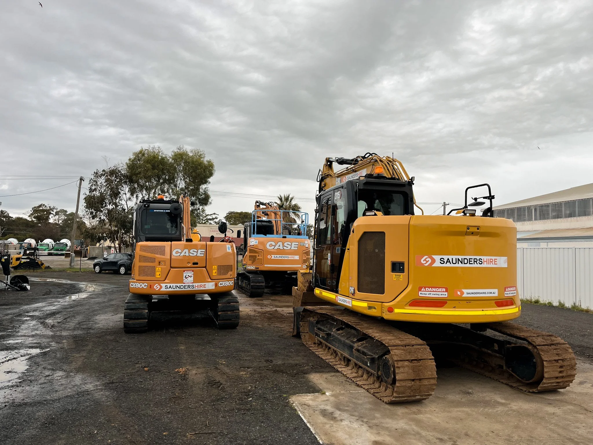 Hire Equipment Machine Western Australia South West WA - Construction yard with yellow excavators and machinery, overcast sky, trees, vehicles, and industrial buildings in background.