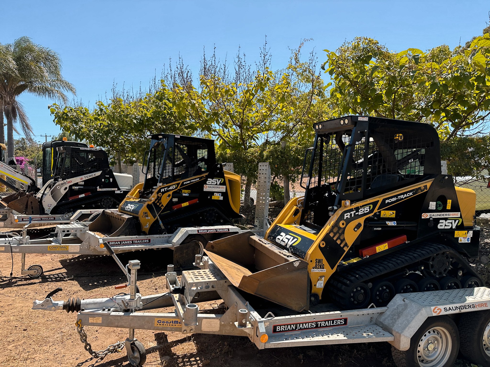 Hire Equipment Machine Western Australia South West WA - Multiple compact construction vehicles, including small bulldozers and trailers, parked outdoors on dirt under a clear blue sky with trees in the background.