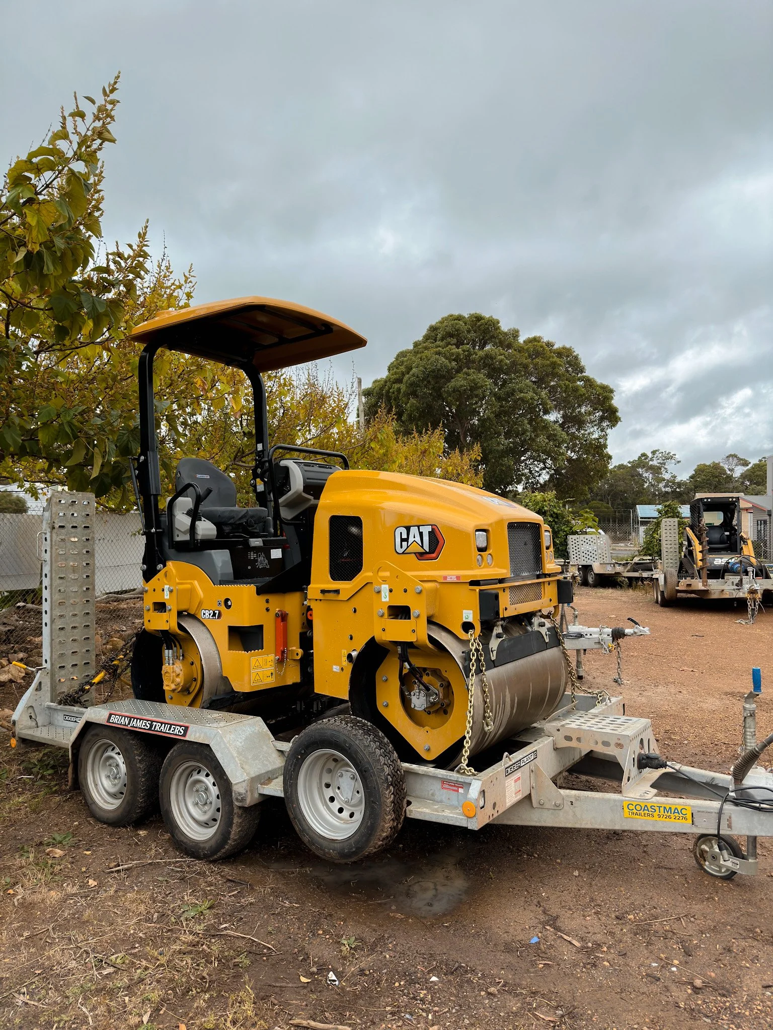 Hire Equipment Machine Western Australia South West WA - A yellow CAT asphalt roller on a trailer at an outdoor construction site, with trees and a cloudy sky in the background.