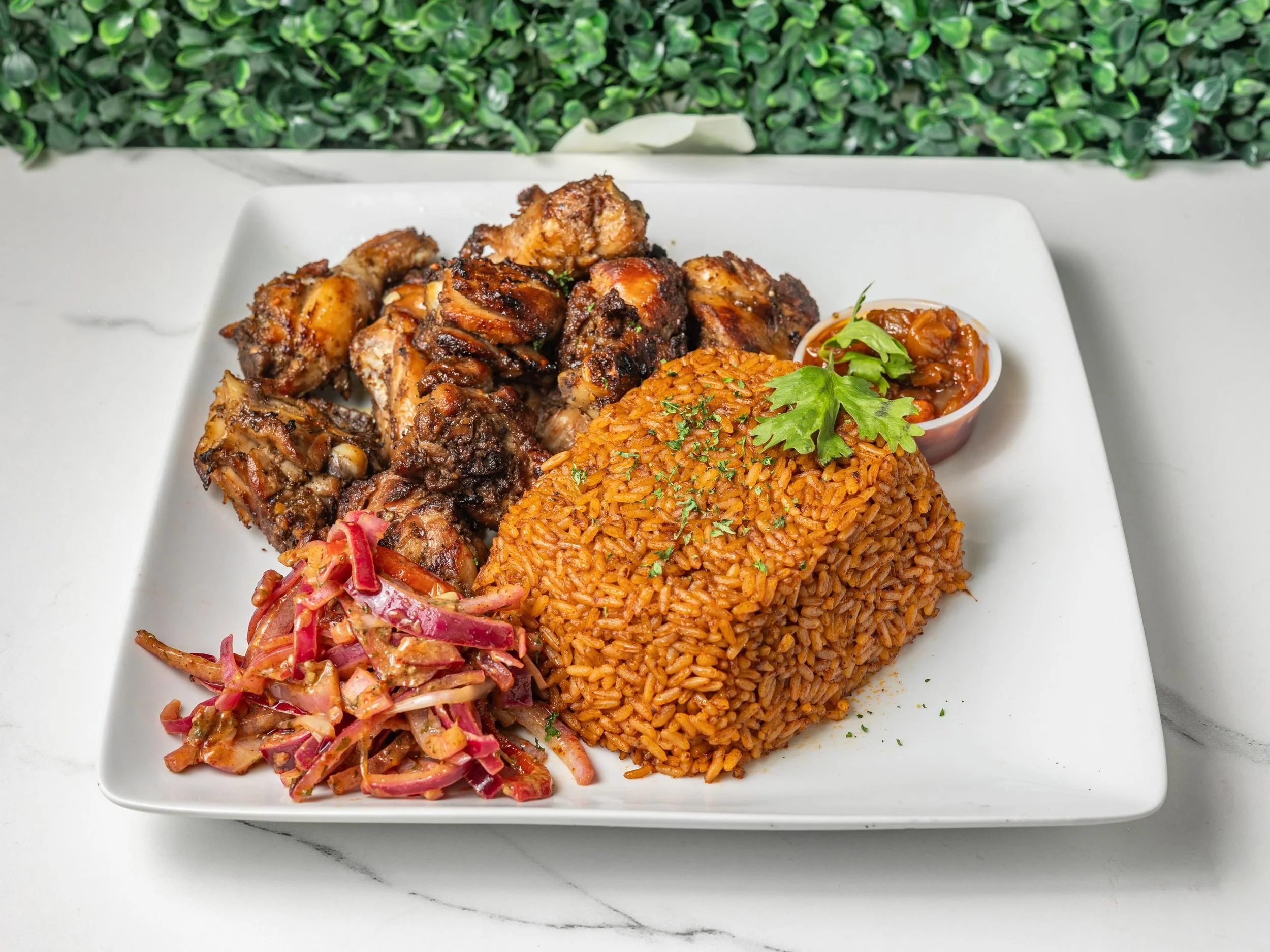 Plate of grilled chicken drumsticks, seasoned rice, and coleslaw garnished with parsley, with a small cup of sauce or stew, on a white rectangular plate.