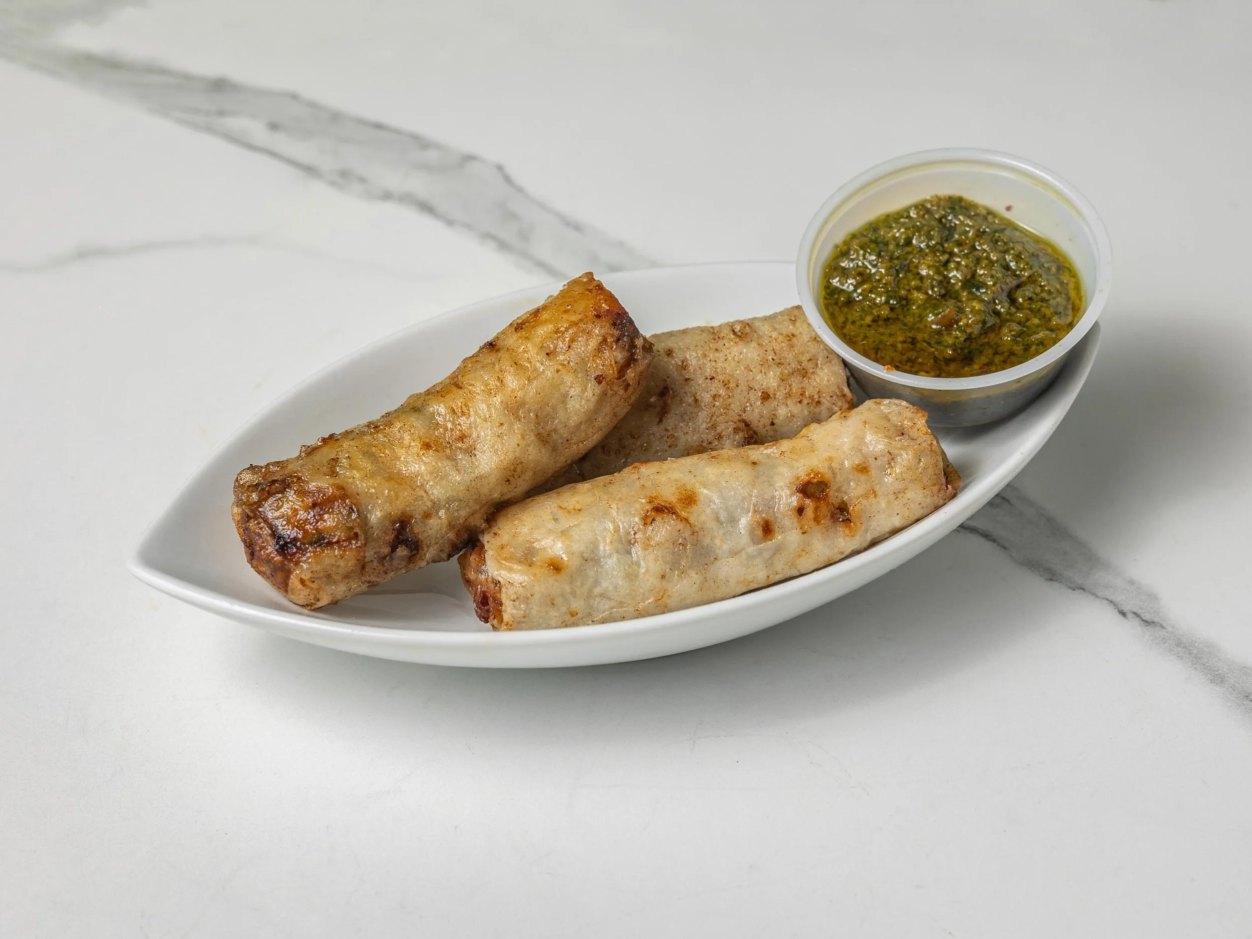 Three spring rolls on a white plate with dipping sauce in a small container, placed on a marble surface.