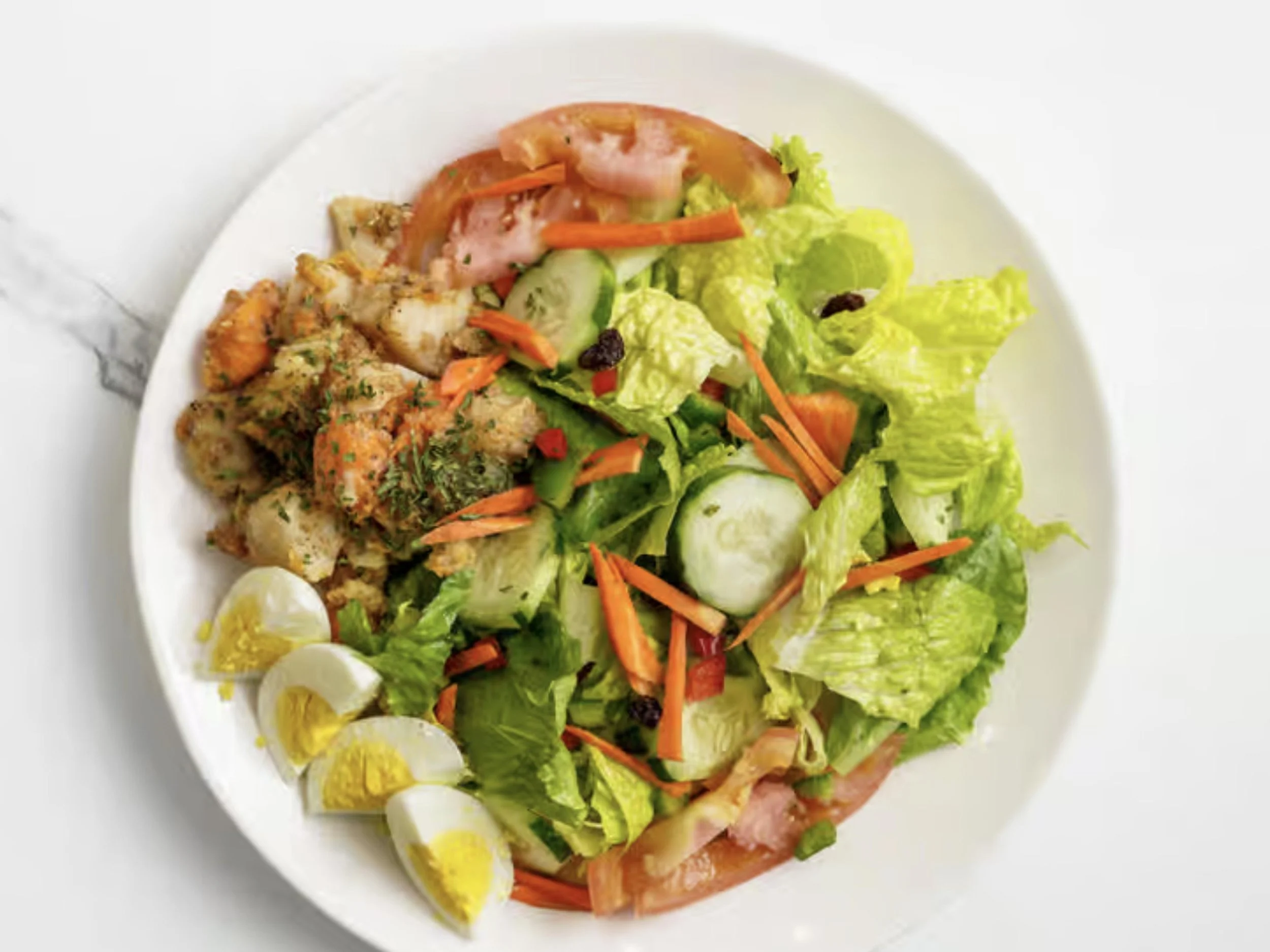 A white plate with assorted salad ingredients including lettuce, cucumber, carrots, cherry tomatoes, sliced lemons, and some cooked chicken pieces garnished with herbs.