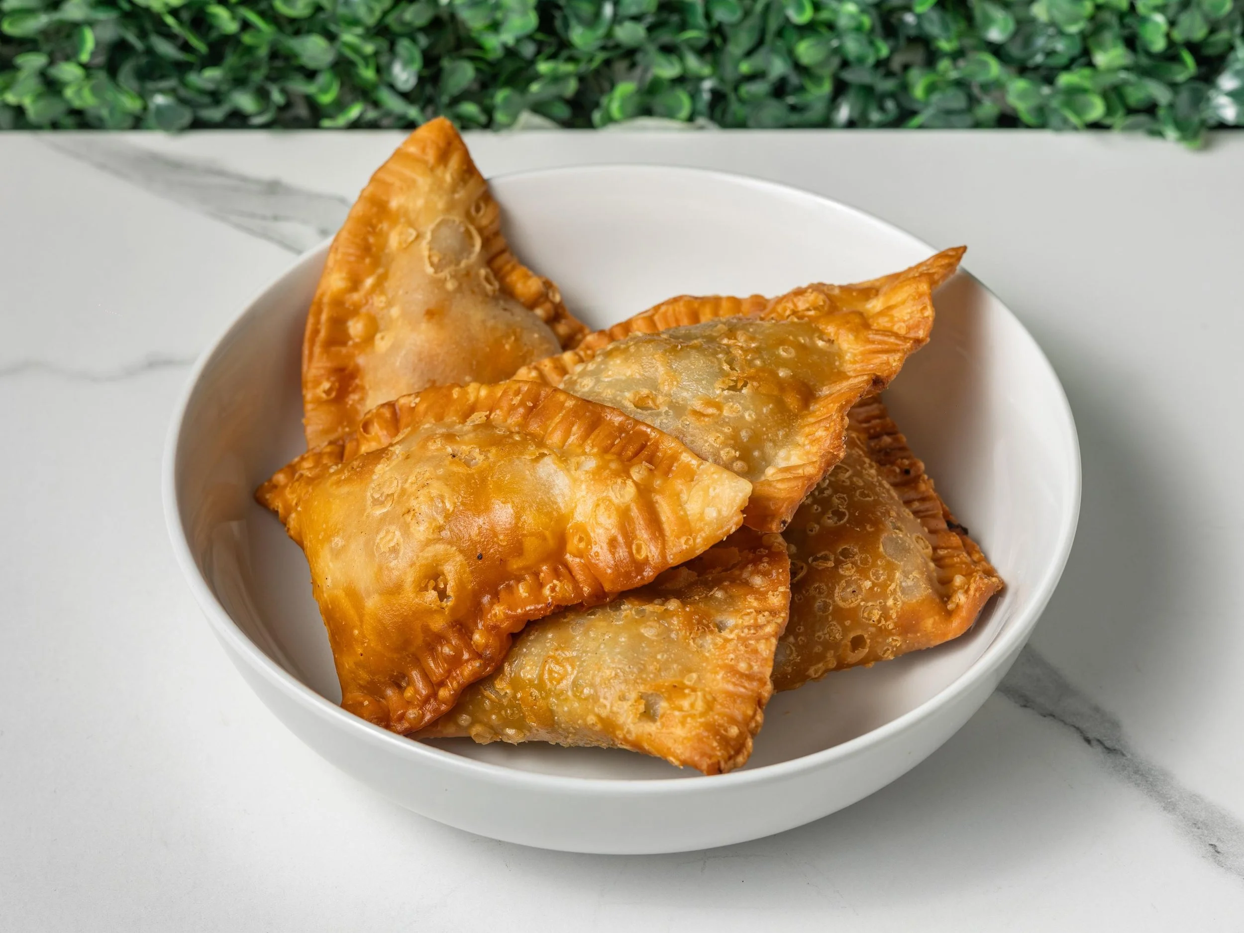 A white bowl filled with fried samosas on a white surface with a green leafy background.
