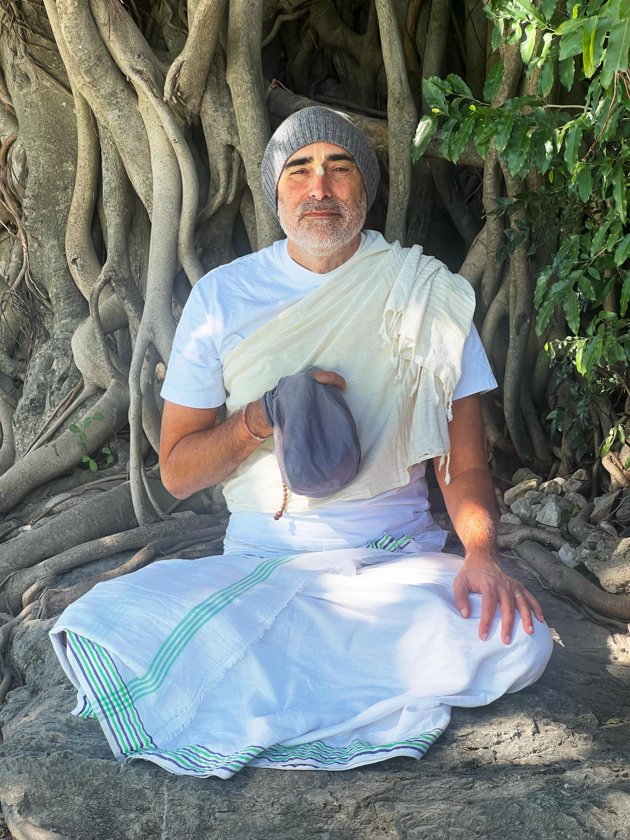 A man sitting outdoors in front of a large tree with roots, dressed in white robes and a gray beanie, with his eyes closed and holding a gray cloth in his hand.