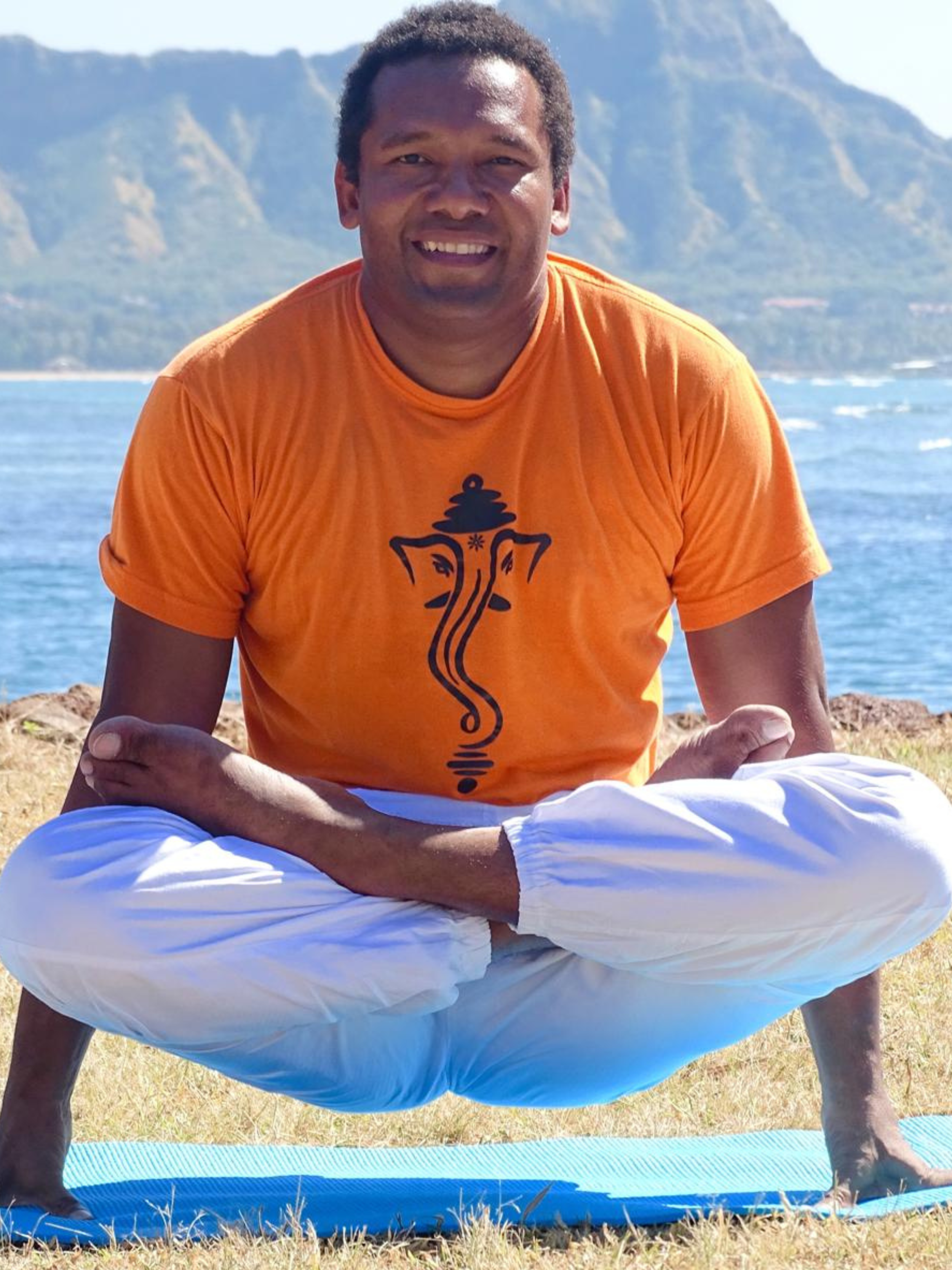 Yogi Charu demonstrating an advanced arm-balance yoga pose outdoors by the ocean, wearing a Ganesh shirt and white yoga pants, with mountains and coastline in the background.