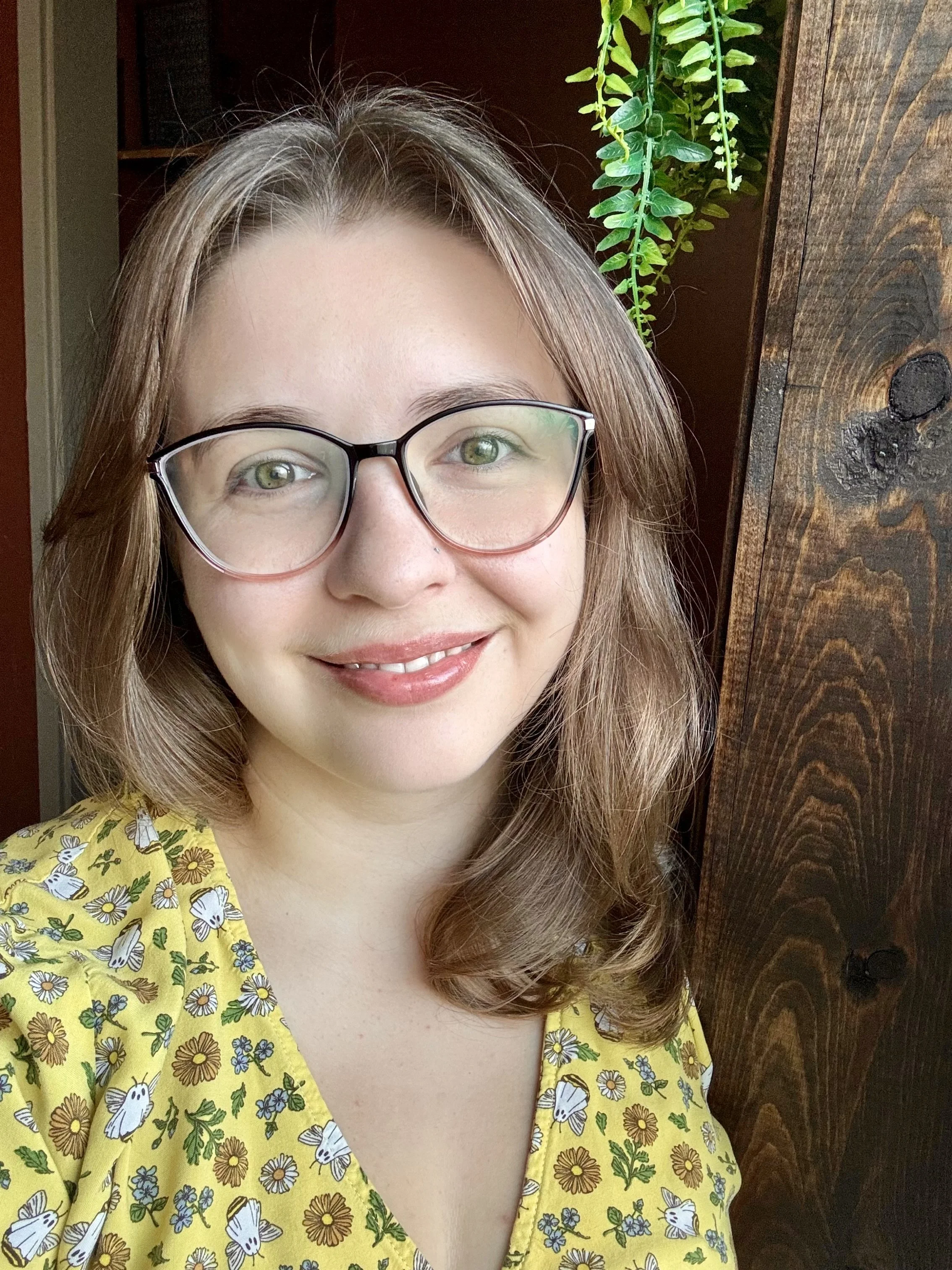 A woman with long, light brown hair and glasses smiling at the camera. She is wearing a yellow dress with a floral and butterfly pattern. Behind her is a dark wooden wall with a green hanging plant.