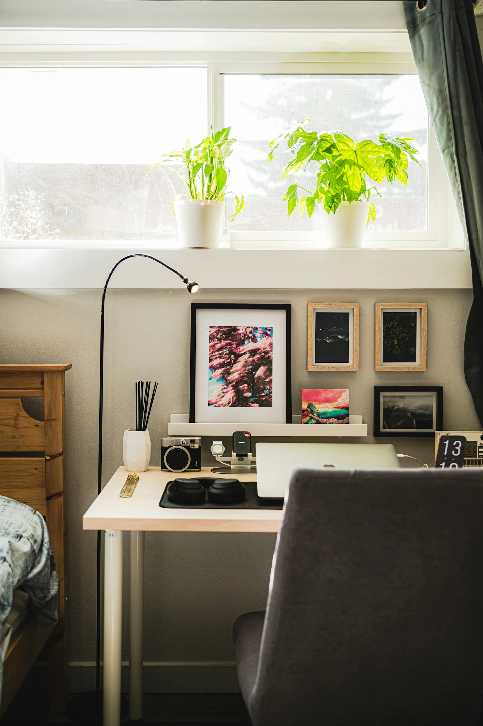 A room with a white desk, a gray chair, and framed wall art, with a window above the desk featuring two potted plants on the windowsill.