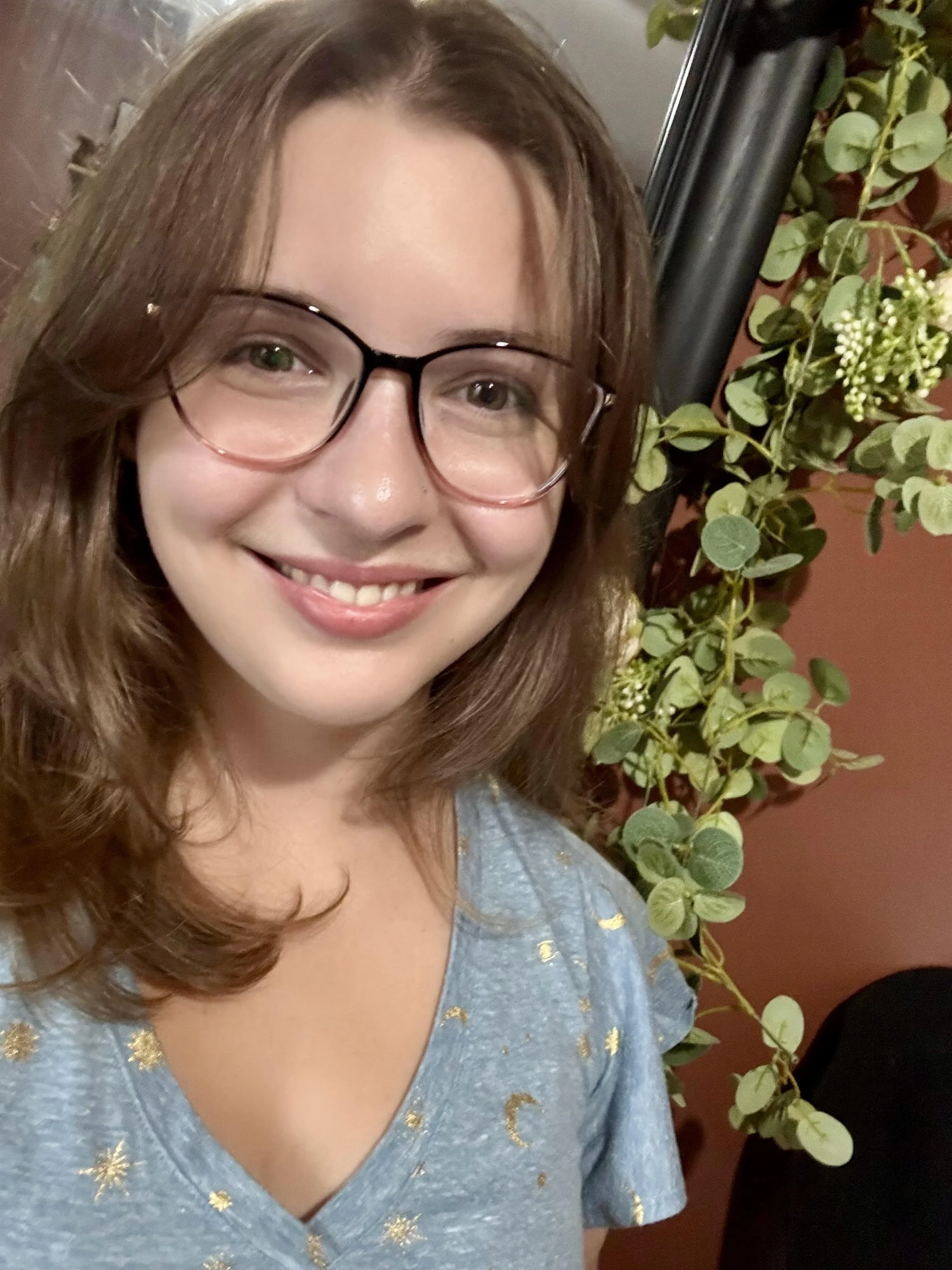 Close-up of a smiling woman with brown wavy hair and glasses, wearing a light blue dress with gold accents, standing beside a green leafy plant against a reddish wall.