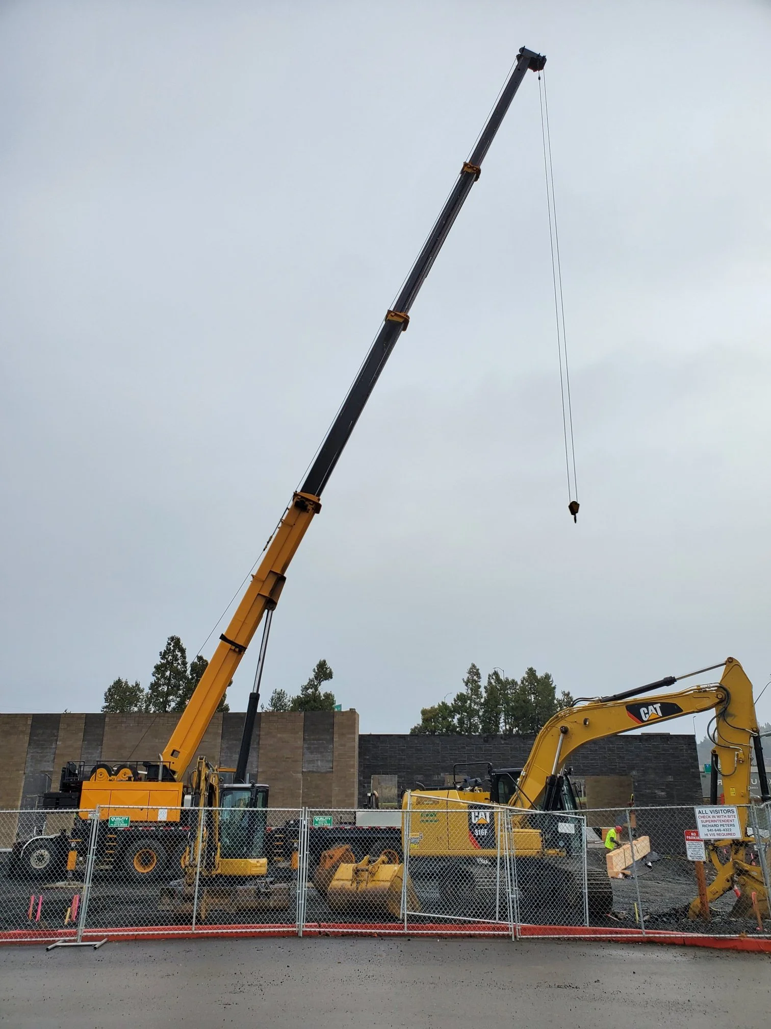 Construction site with a large yellow and black crane and excavator, surrounded by a chain-link fence with construction signs, and trees in the background under an overcast sky.