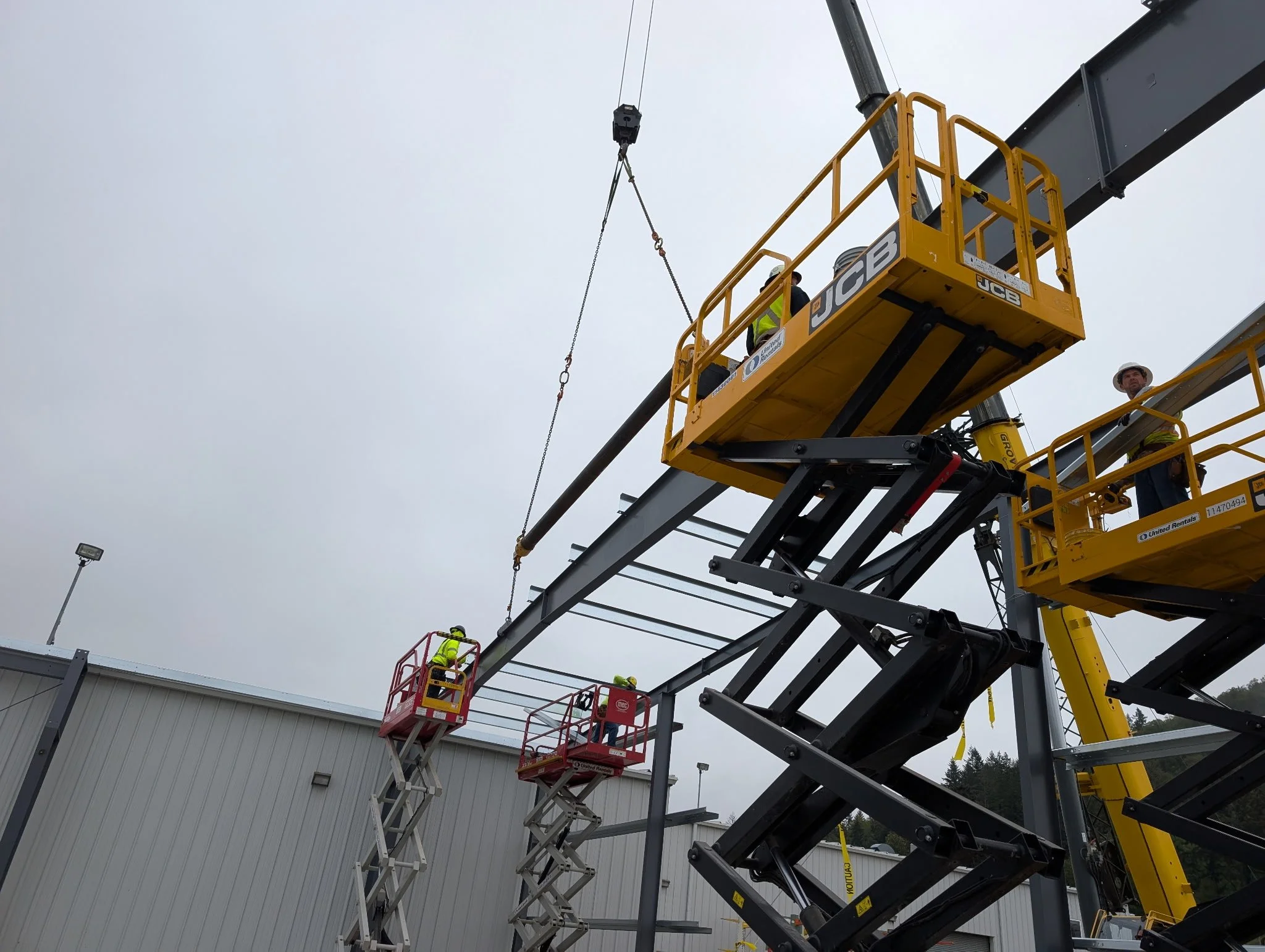 Construction workers in safety vests and hard hats working on a large outdoor steel framework using scissor lifts and a cherry picker under an overcast sky.