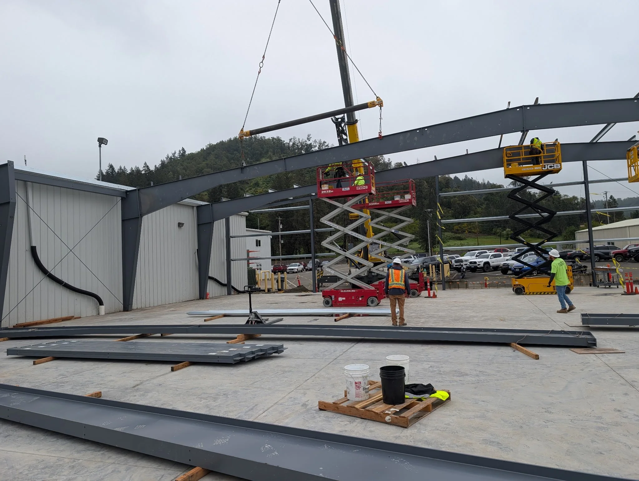 Construction crew installing structural steel roof beam inside commercial building frame using crane and elevated work platforms