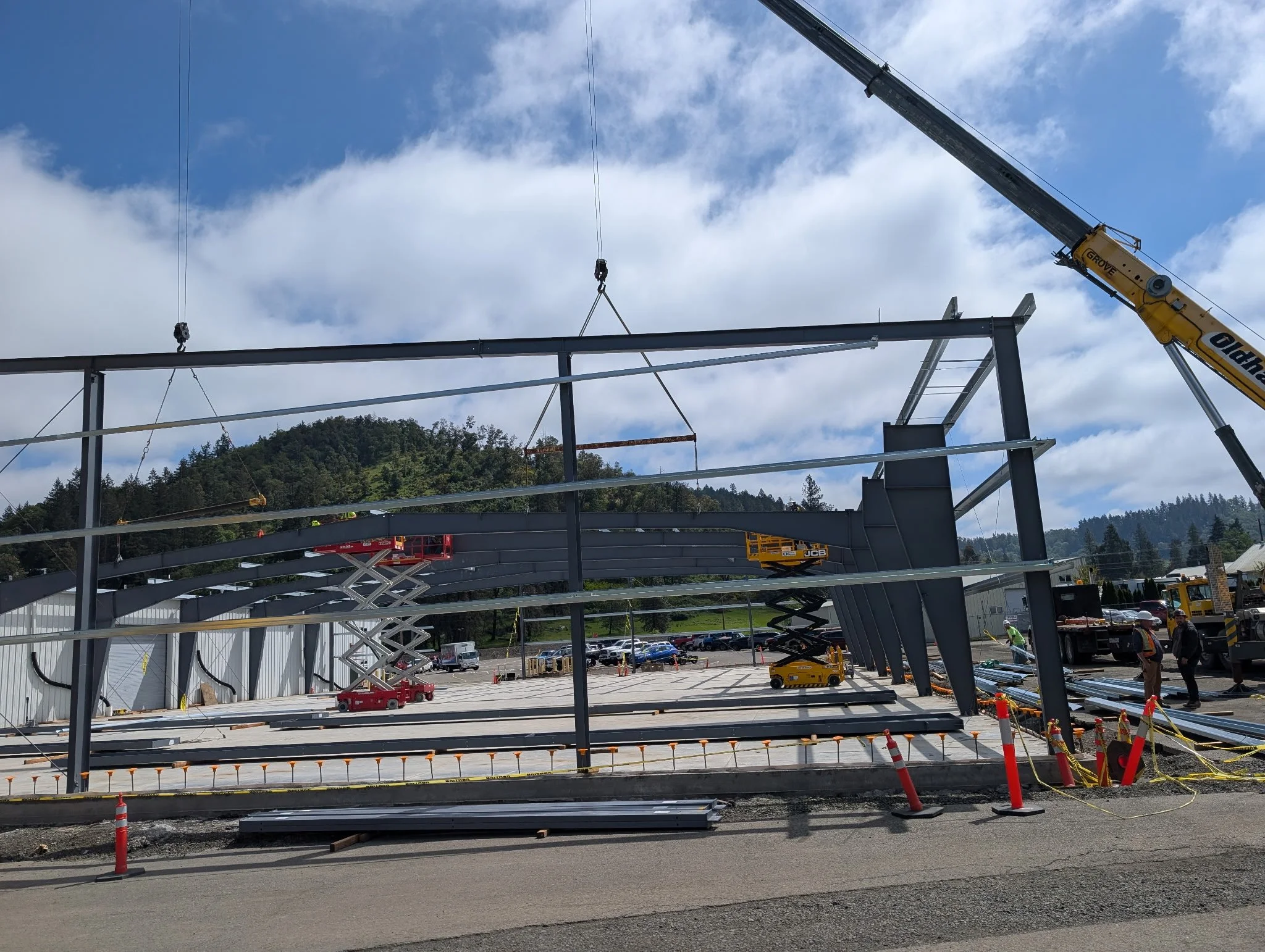 Construction site showing steel framework being assembled, with cranes and construction workers. Hills with trees in the background under partly cloudy sky.
