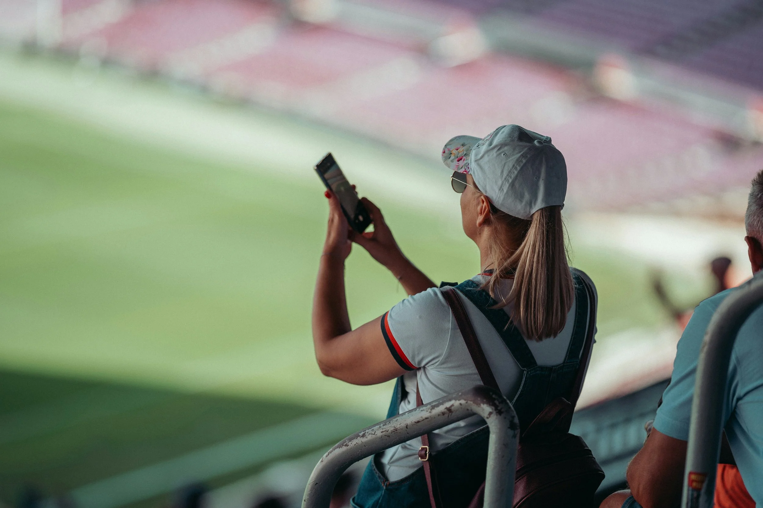A woman wearing a light grey cap, sunglasses, and a white shirt with red and black accents is sitting on a stadium seat. She is holding a phone in her right hand and appears to be looking at it. The background shows other stadium spectators and a blurry green field.