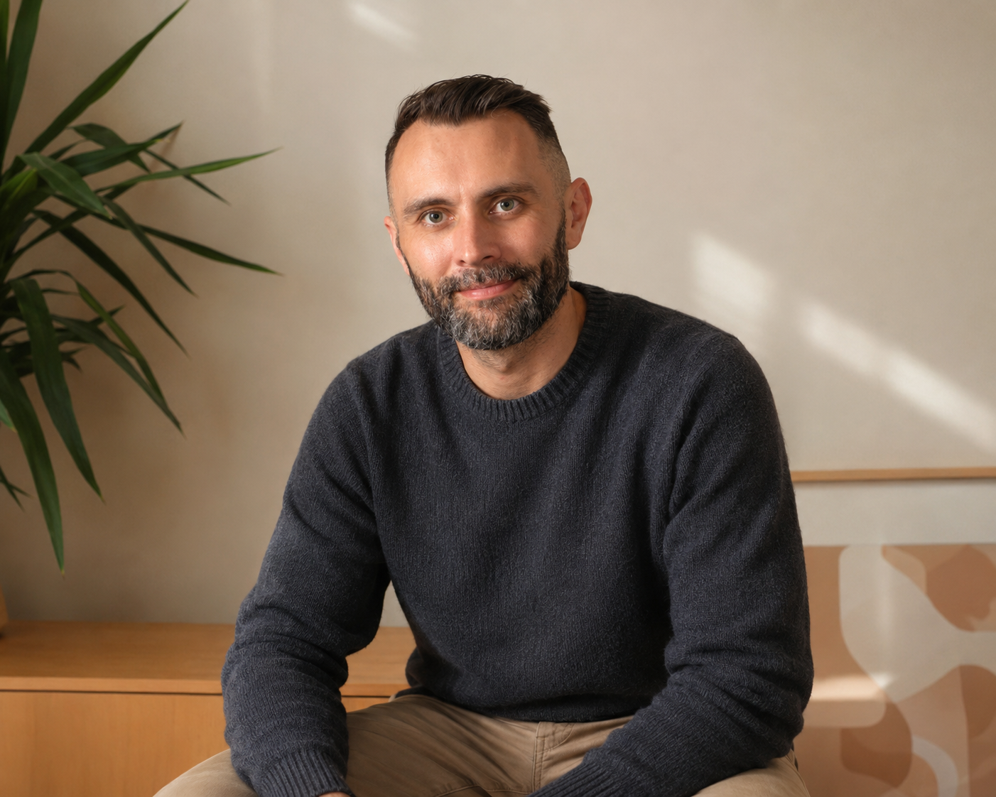 Man with short dark hair and beard wearing a dark gray sweater, sitting on a wooden bench, with a large green plant and a beige wall in the background.