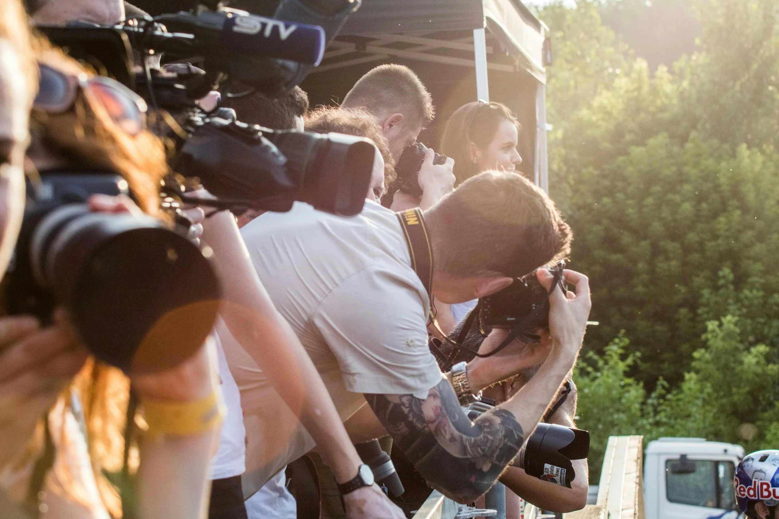 Group of people with cameras and binoculars at an outdoor event likely during sunset with trees in the background.