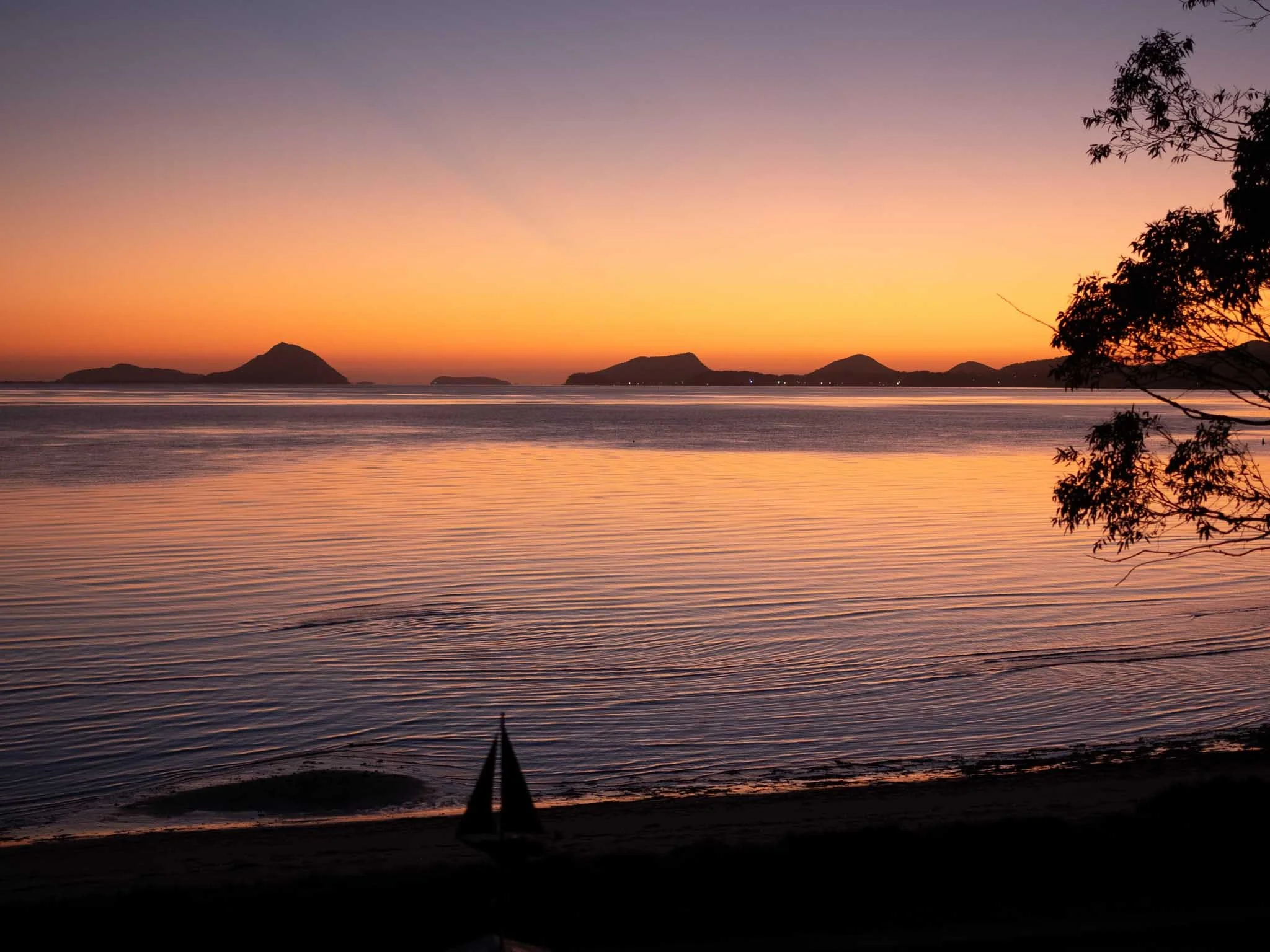 A sunset over calm water with distant islands and a silhouette of a tree on the right side of the image.