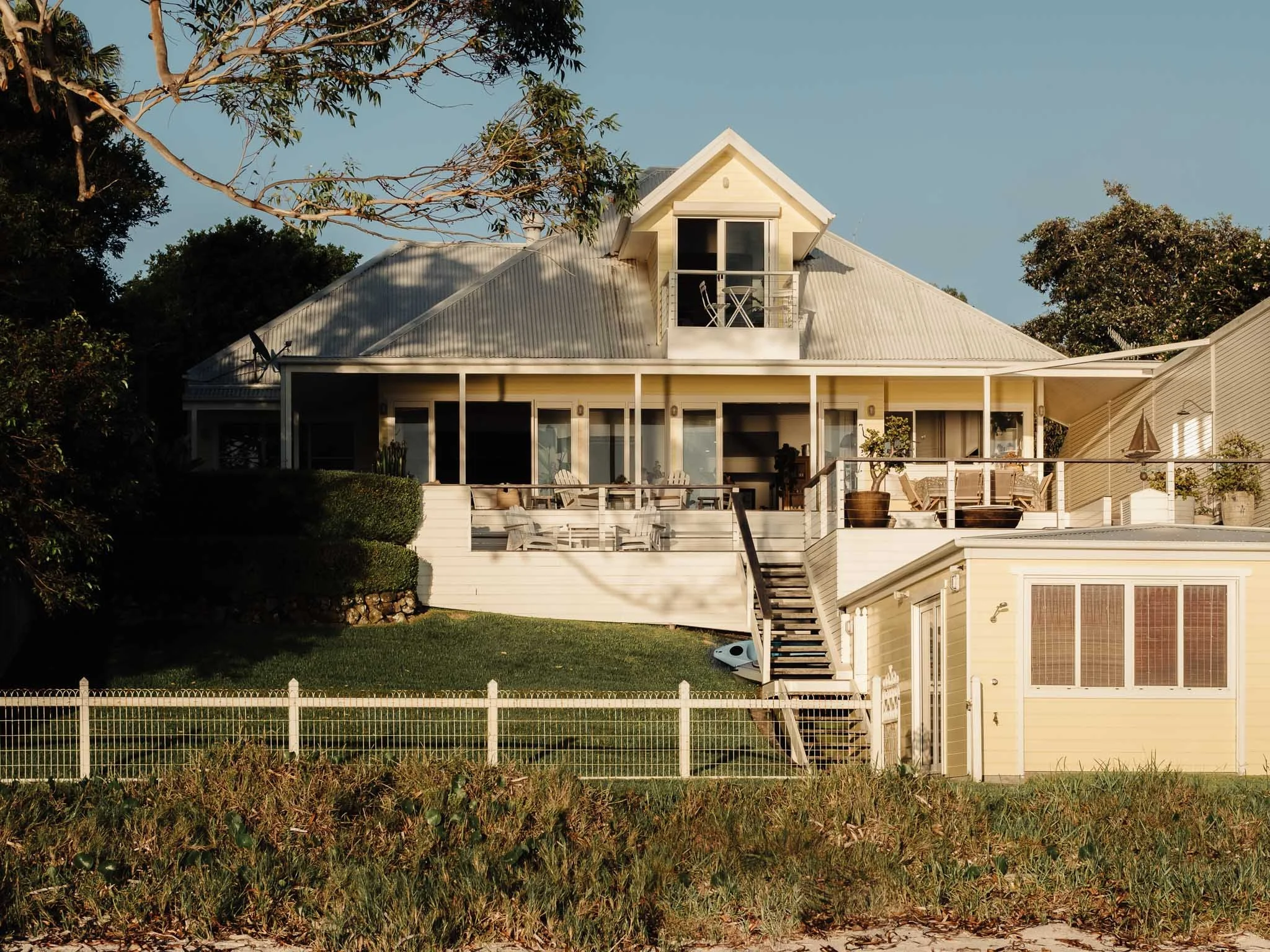 A multi-story house with a white exterior, large windows, a small balcony, and outdoor furniture. The house has a corrugated metal roof and a staircase leading to a second-floor deck. The property is enclosed by a white picket fence, with trees and greenery surrounding it.
