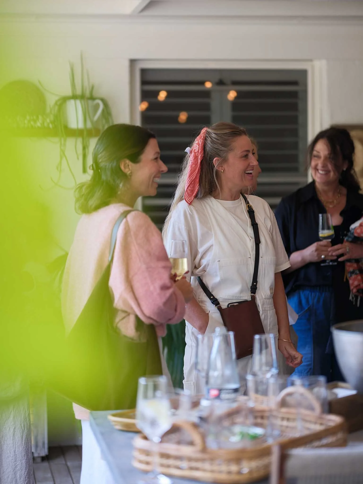 Three women laugh and converse at a social gathering indoors. There are wine glasses on a table with baskets in the foreground, and a window with blinds behind them.