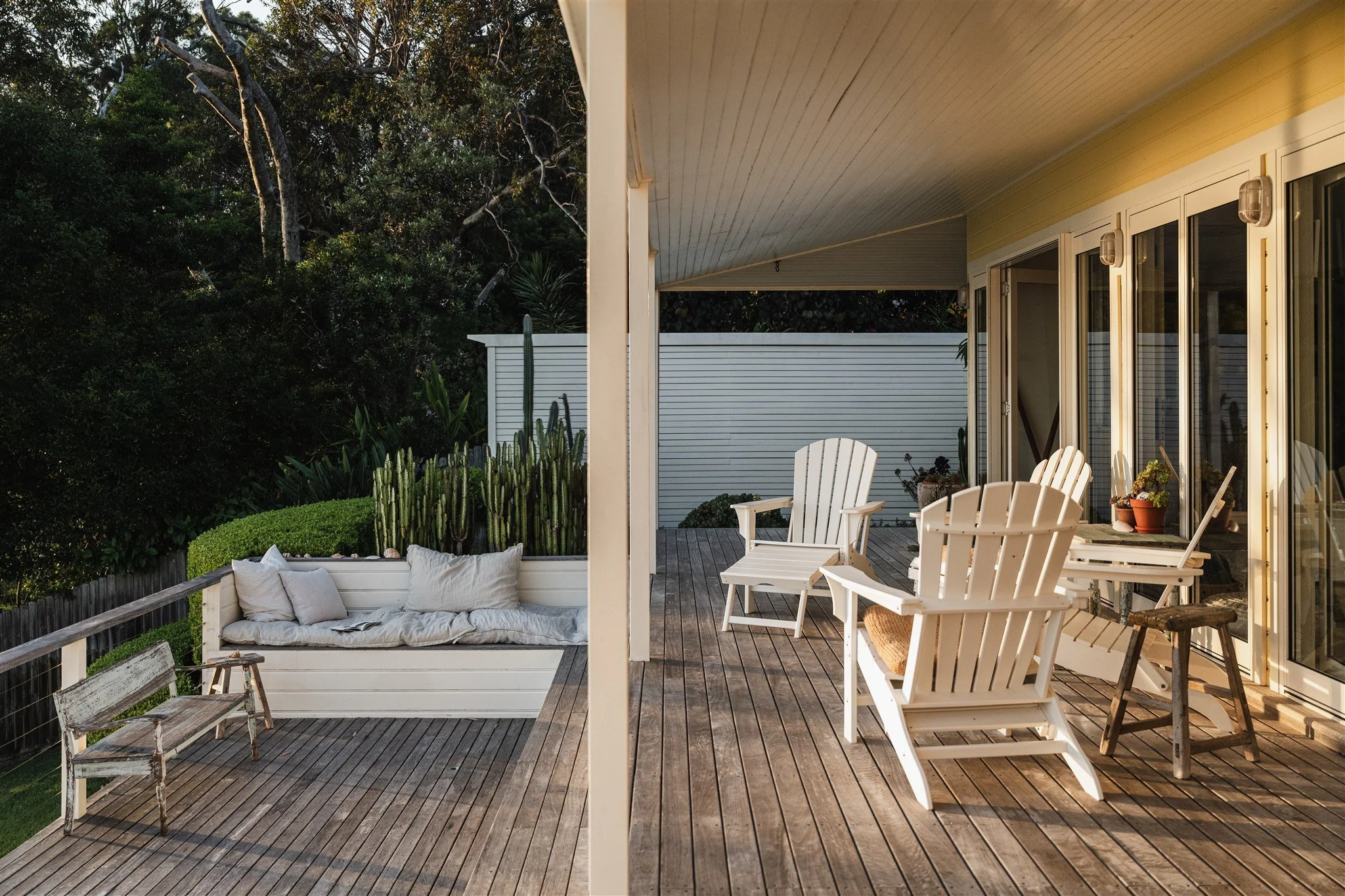 A split view of an outdoor patio showing on the left a white cushioned outdoor sofa with pillows and an old wooden chair, and on the right a dining area with white wooden chairs, a small side table, and potted plants, with a background of lush green trees.