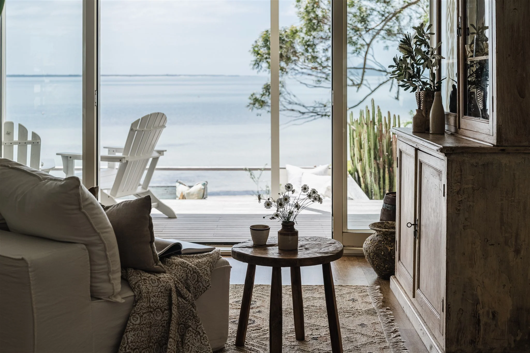Living room with large glass sliding doors opening to a beach view, with white chairs outside, and cozy furniture and plant decor inside.
