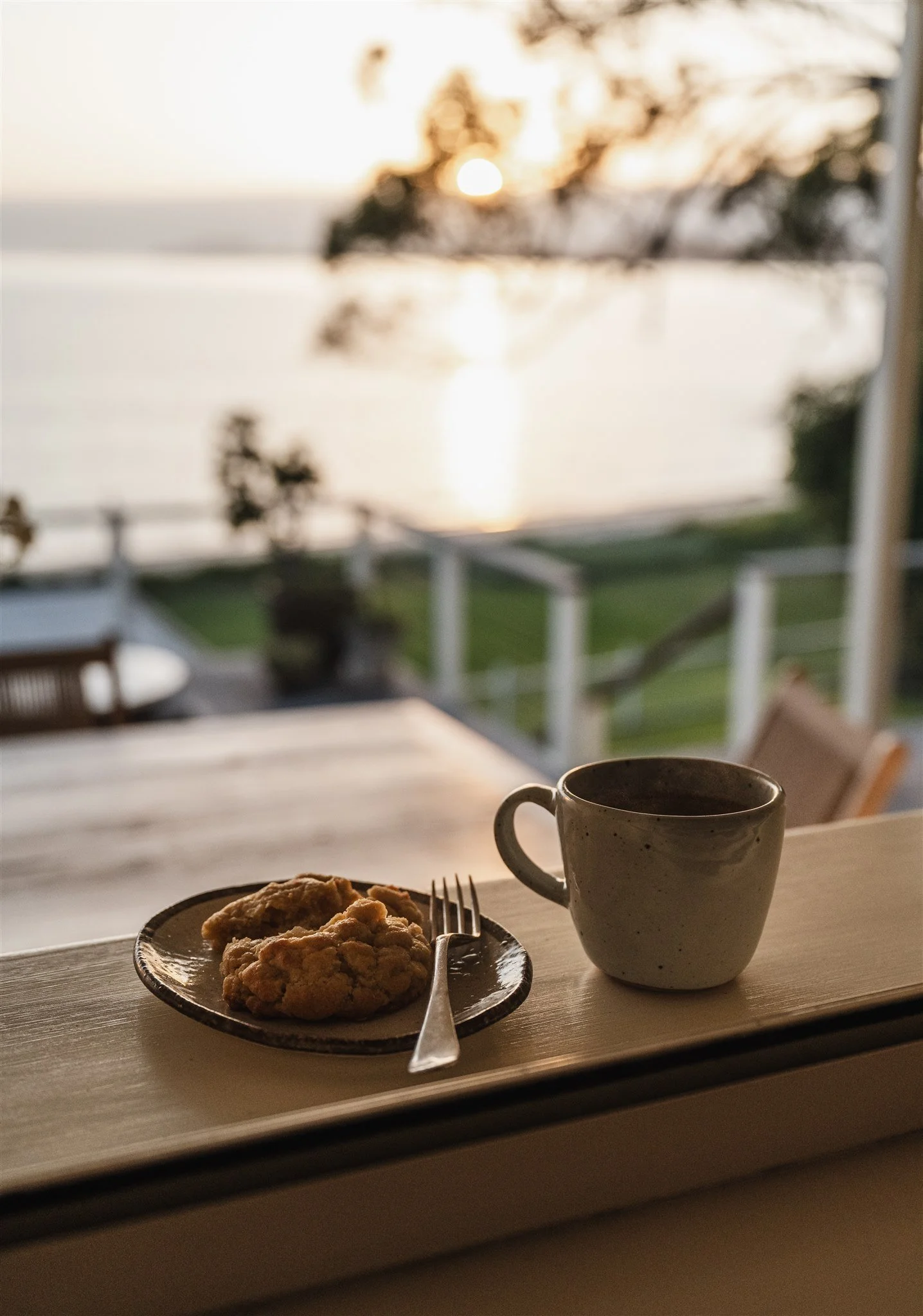 A breakfast scene with cookies on a plate, a white ceramic mug, a fork, and a window view of a sunset over the water with trees.