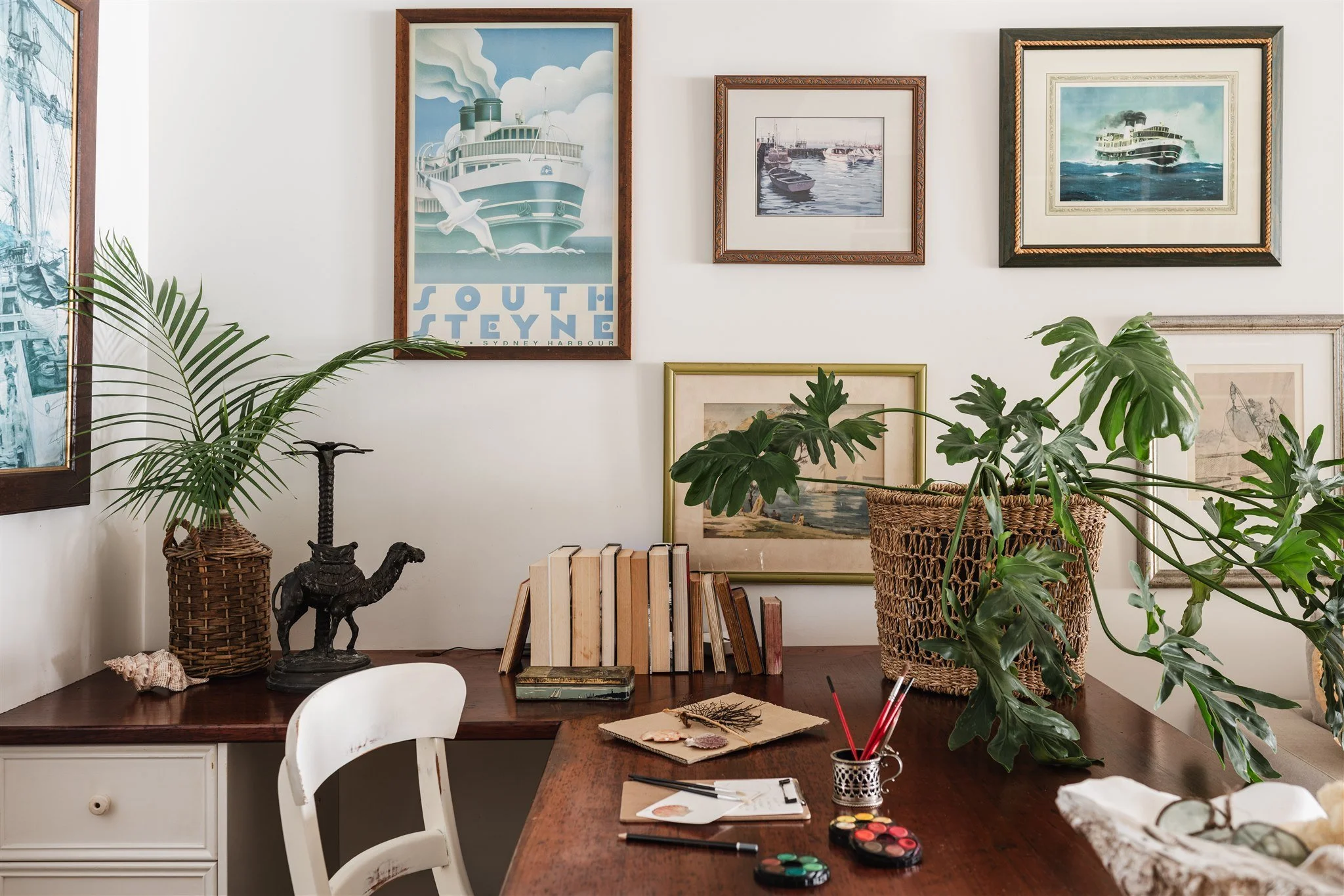 A cozy home office with framed maritime posters on the wall, large leafy plants in wicker baskets, and a cluttered wooden desk with art supplies, books, and decorative objects.