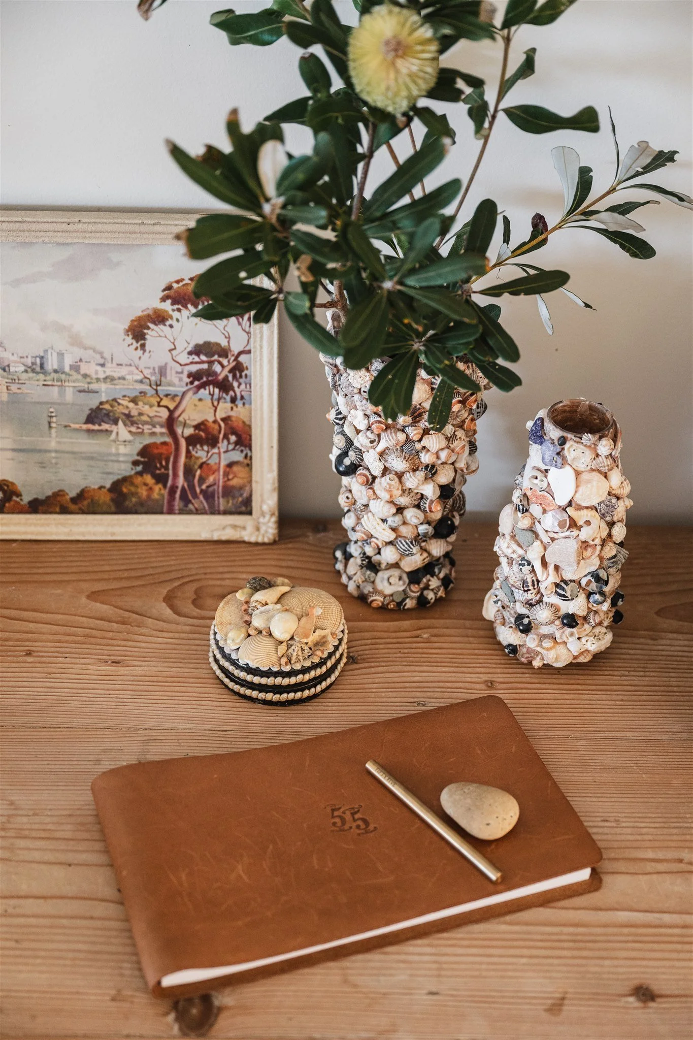 A wooden desk with a brown journal, a pen, a shell, and a small stone, with decorative shell vases and a framed picture of a landscape on the wall behind.