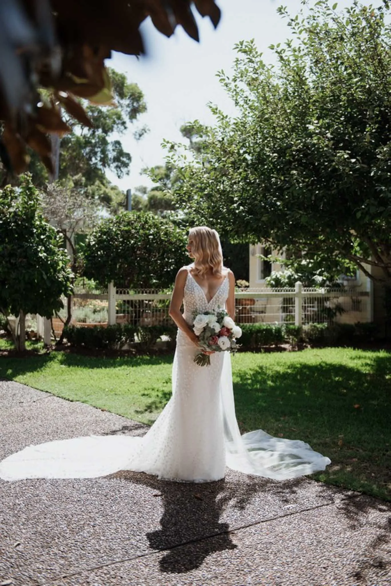 A woman in a white wedding dress stands on a path outside, holding a bouquet of flowers, with lush green trees and a white fence in the background.