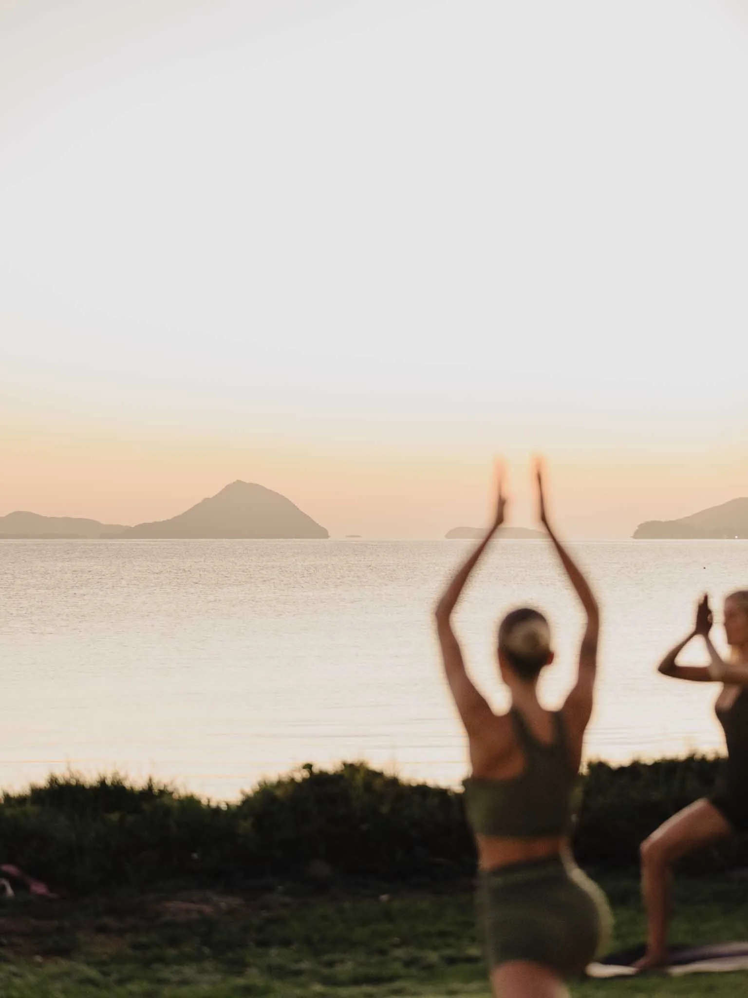 Two people practicing yoga or meditation outdoors near a body of water during sunset or sunrise. The scene includes silhouettes of mountains or islands in the background.
