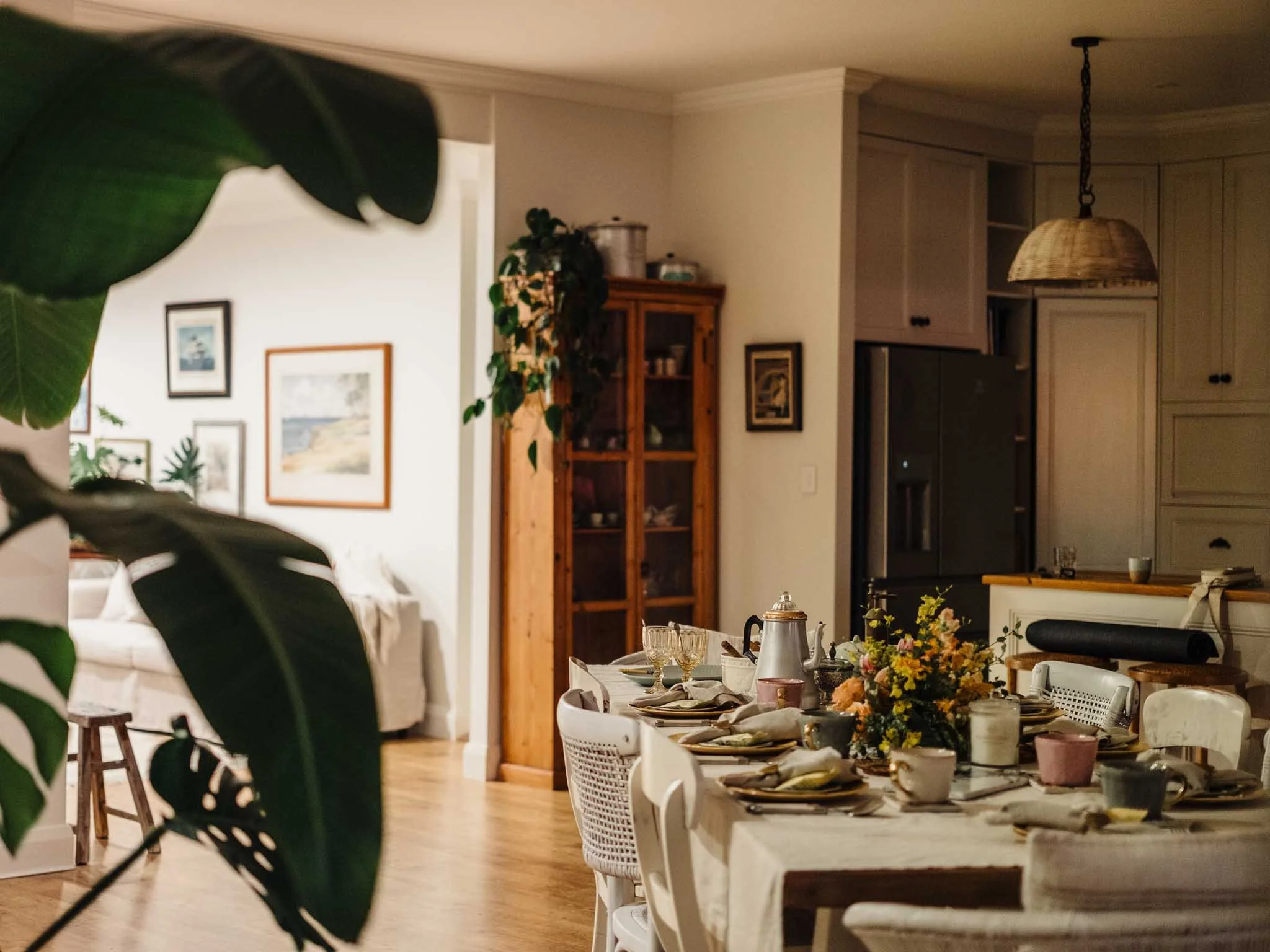 A dining area set for a meal with plates, cups, silverware, and a floral centerpiece on a long table, with a kitchen and wall art in the background.