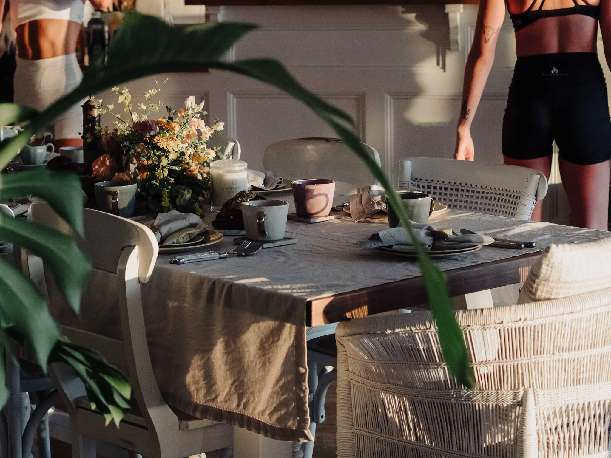 A dining table set with floral centerpiece, cups, plates, napkins, and cutlery in a bright room with wicker chairs. Partially visible people are standing in the background.