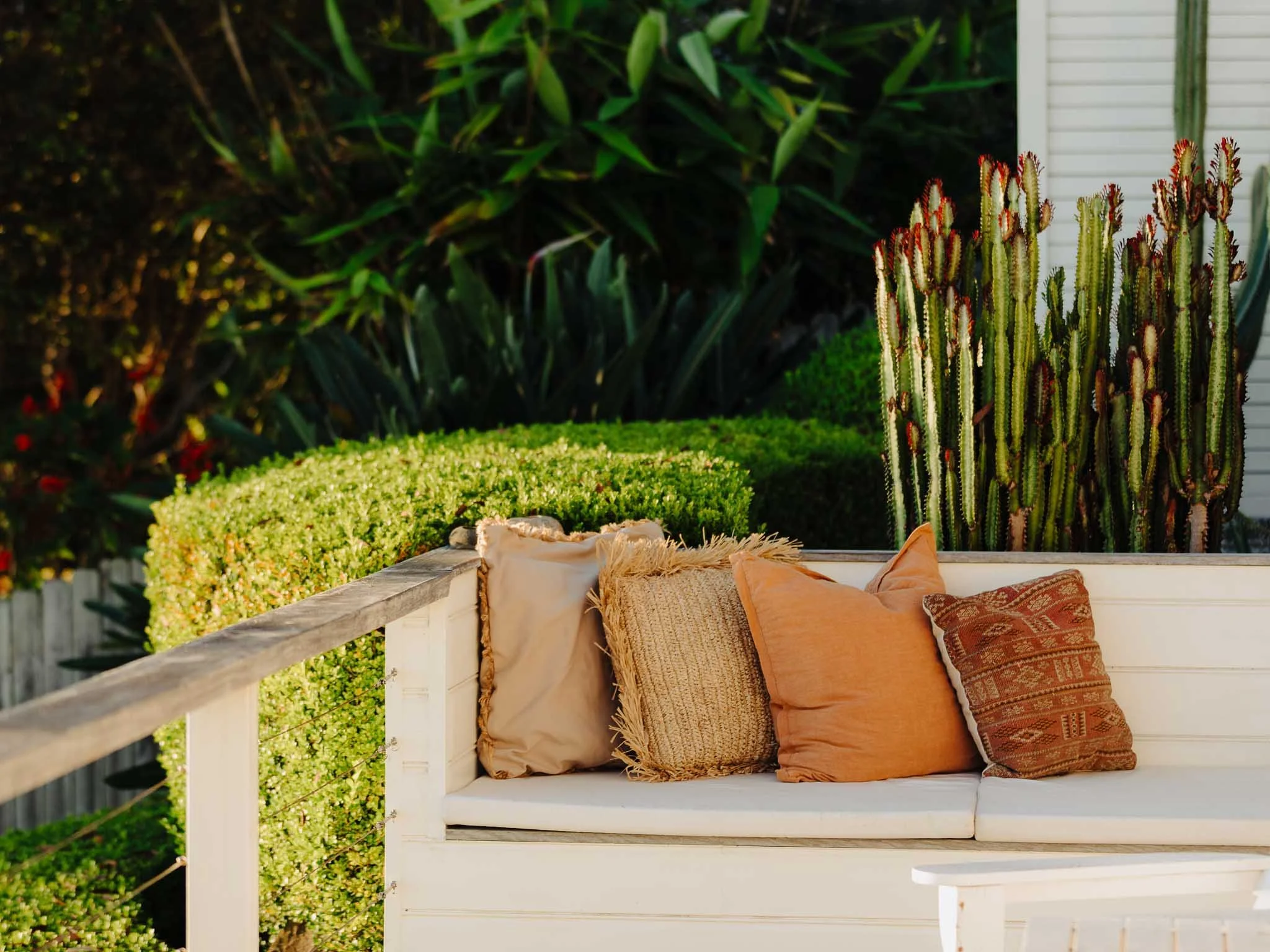 Outdoor patio with a white bench adorned with beige, orange, and patterned cushions, surrounded by lush green plants and a white house siding in the background.