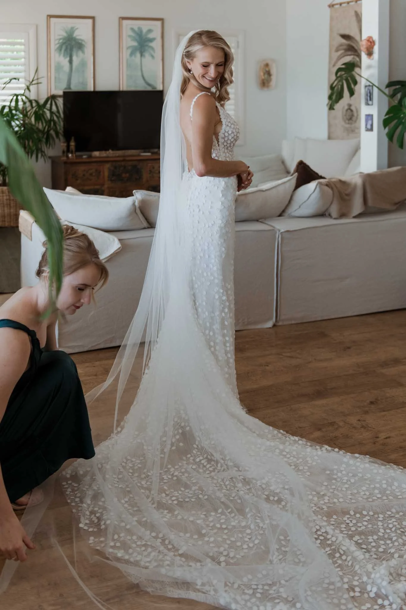 A bride in a white wedding gown with a long train, standing in a cozy living room, smiling and looking down, as a woman adjusts her dress. The room has a beige couch, green plants, and wall art featuring palm trees.