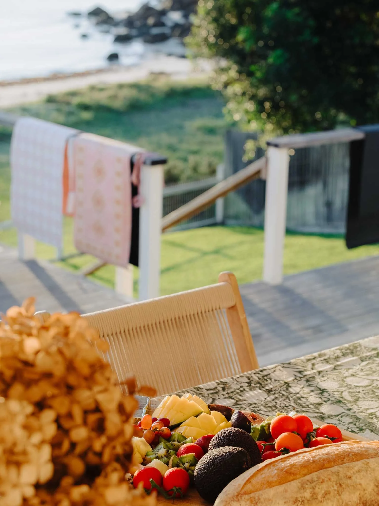 A table set with a variety of cheeses, cherry tomatoes, and fruits, with a scenic view of a backyard and a body of water in the background.