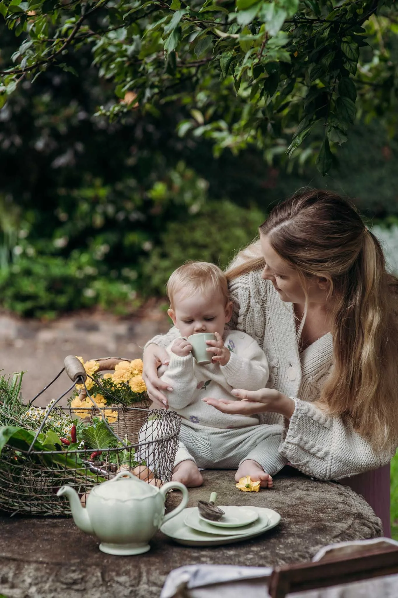 A woman and a young child sitting at a rustic outdoor table with a basket of yellow flowers, teapot, and tea set, surrounded by greenery and trees.