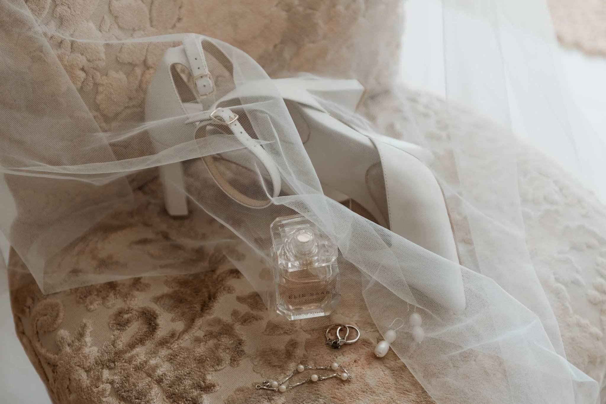 Close-up of a white high-heeled shoe with a satin finish, placed on a marble surface. A sheer white veil drapes over the shoe, with pearl and metallic jewelry pieces also on the surface.