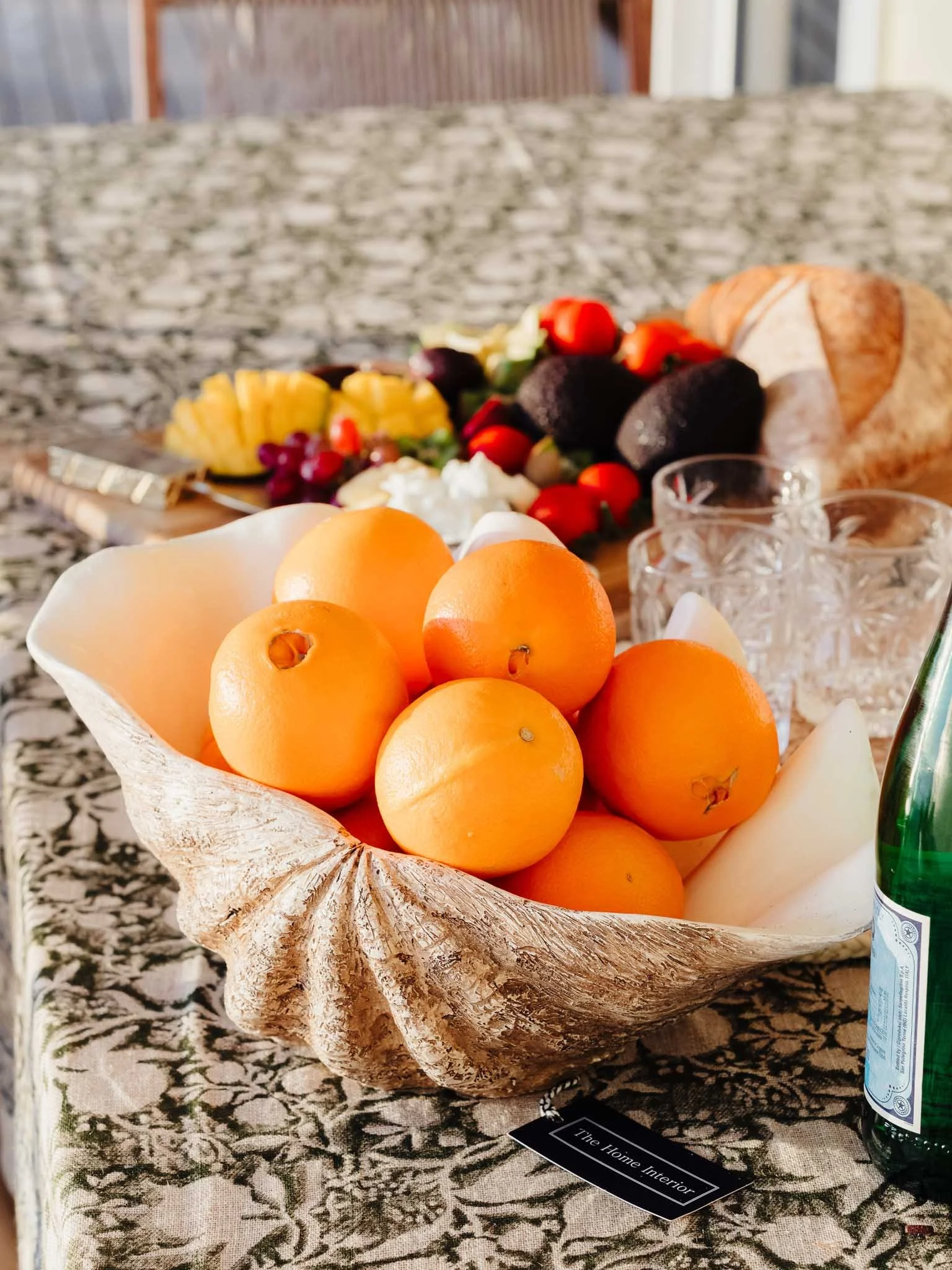 A bowl of oranges on a table with a decorative interior, with a platter of assorted fruit and bread in the background.