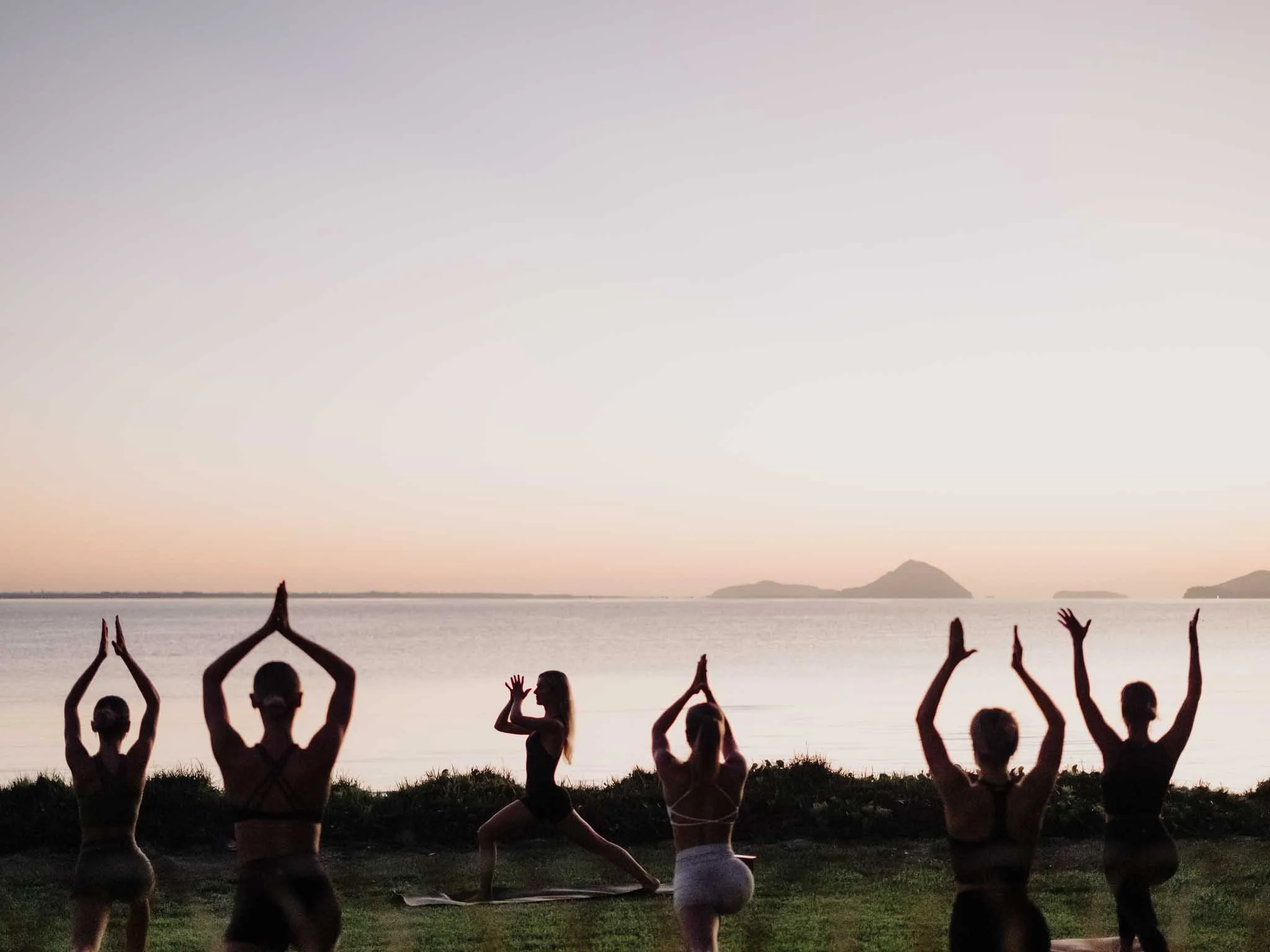 Silhouettes of people practicing yoga on a beach at sunset, facing the water with mountains in the distance.