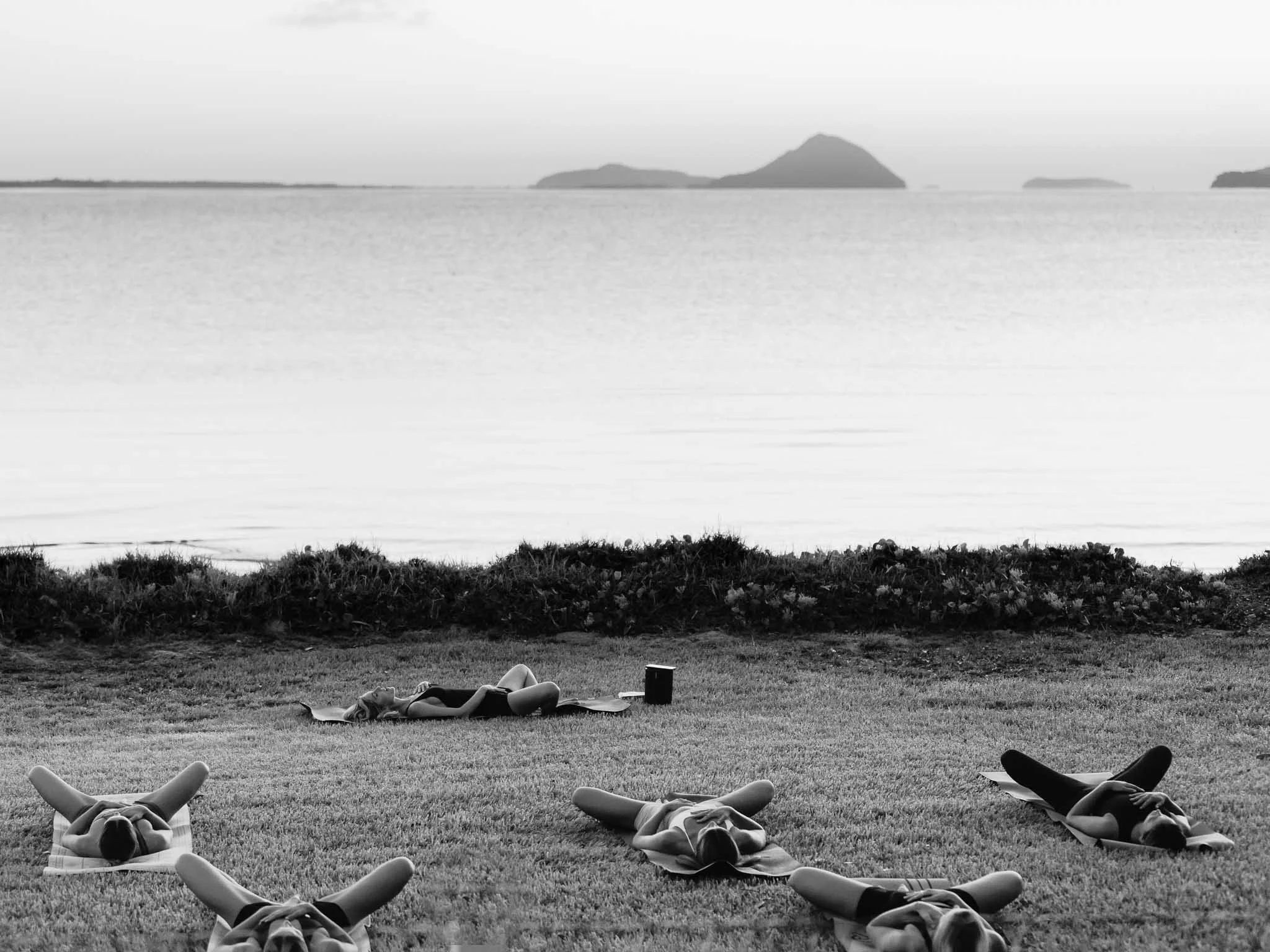 People practicing yoga outdoors on a grassy field near a body of water with islands in the background.