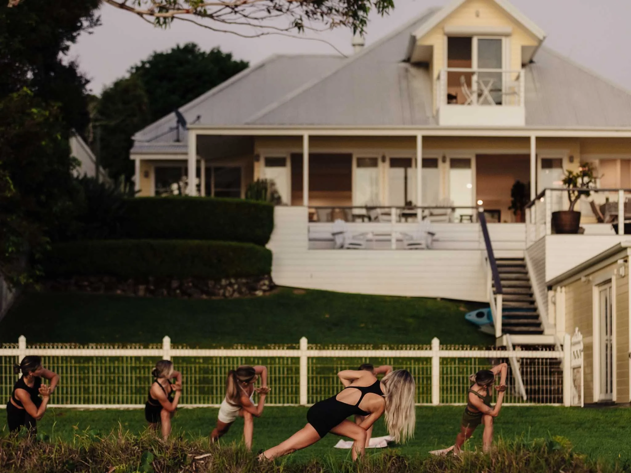 Women practicing yoga outdoors on a grassy area behind a white fence, with a yellow house with a balcony and a porch in the background, trees, and animals in the yard.