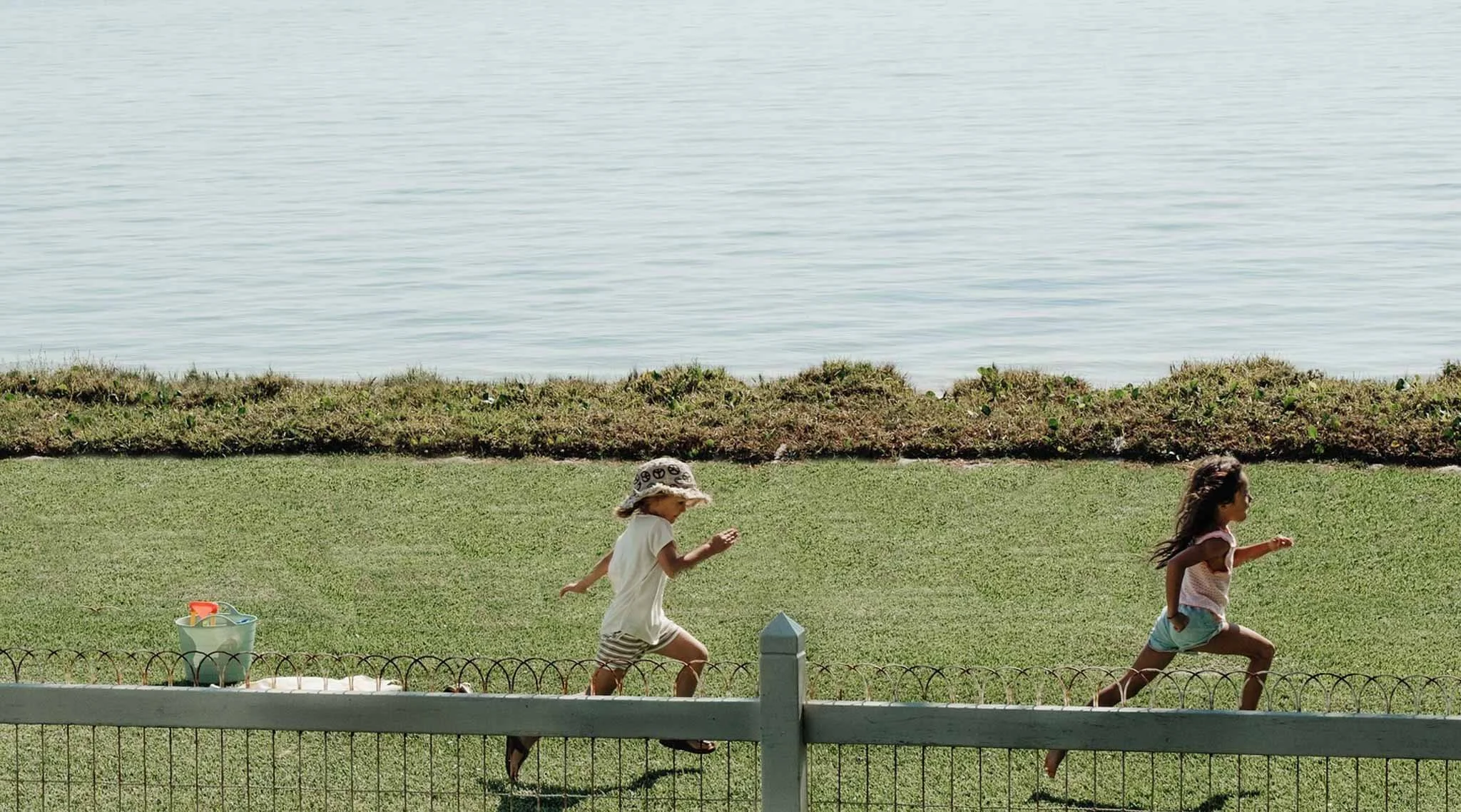 Two children running on a grassy lawn near a body of water, with a bucket and toy shovel on the ground and a white picket fence in the foreground.