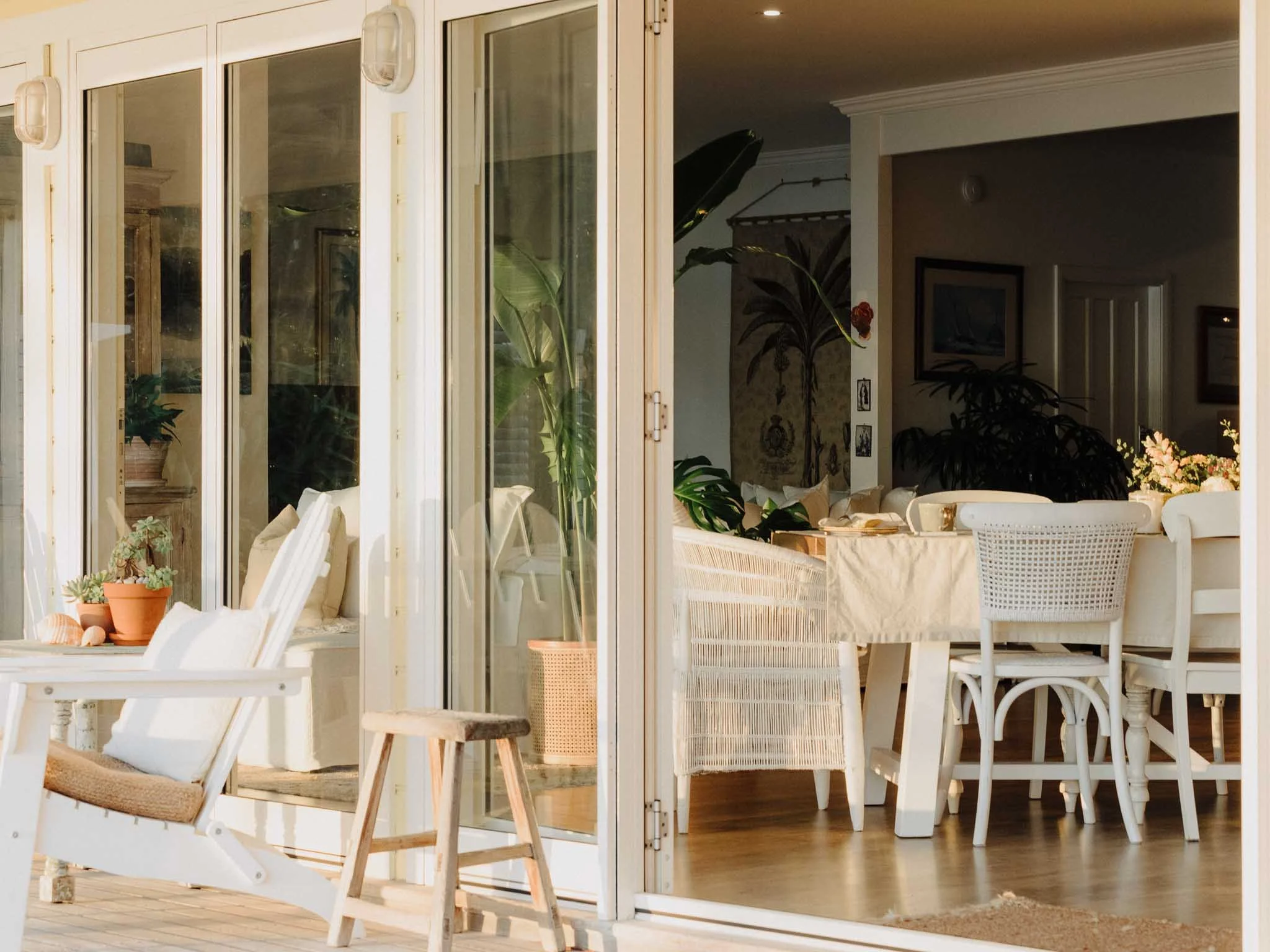 A bright, airy indoor dining area with white furniture, large glass sliding doors, and various indoor plants. There is a table with chairs, a small wooden stool, and sunlight illuminating the space.
