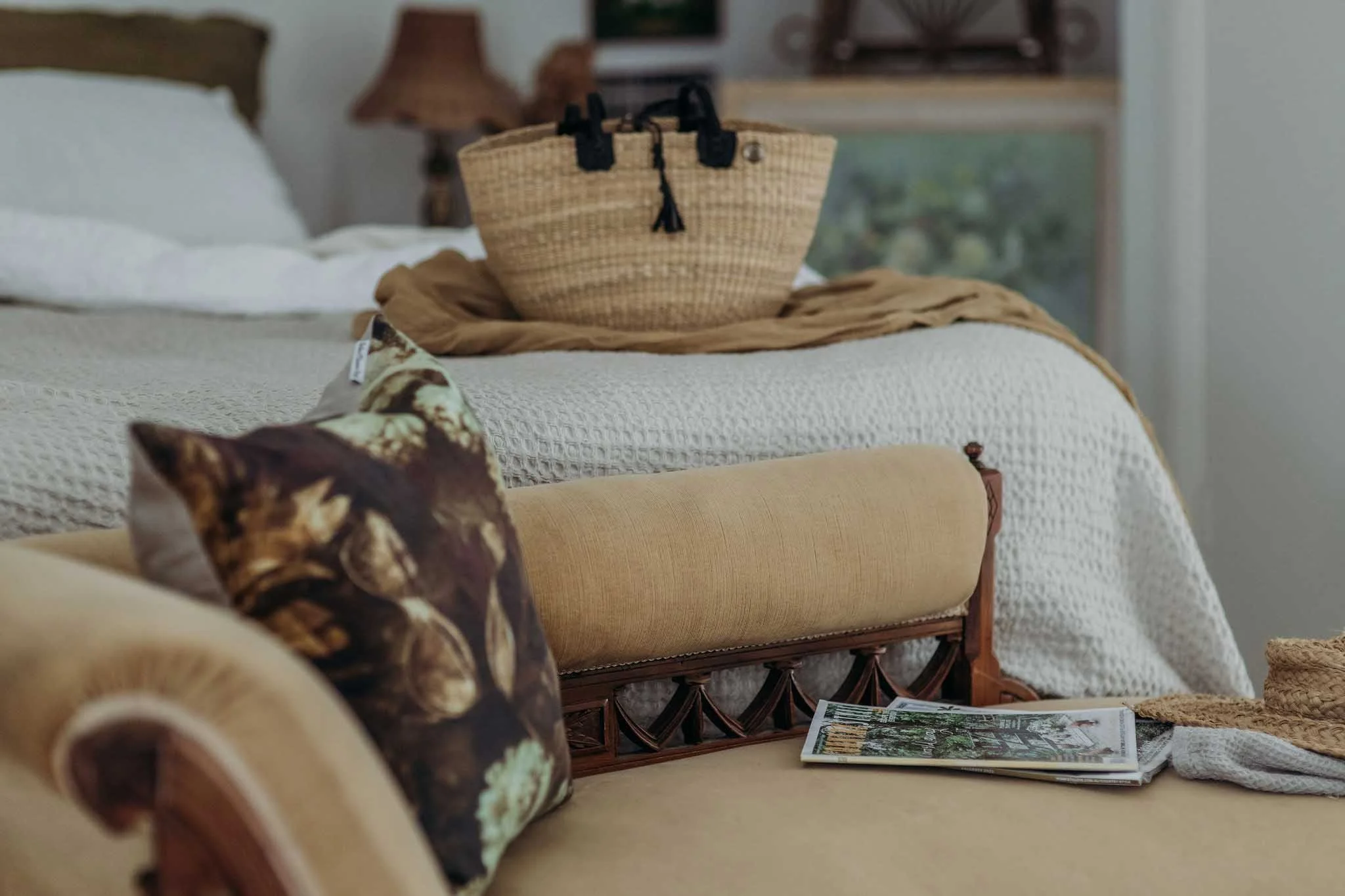 A cozy bedroom scene featuring a bed with white bedding, a wicker handbag on a bedspread, a brown armchair with floral upholstery, a newspaper on the beige ottoman, and a window with greenery outside.